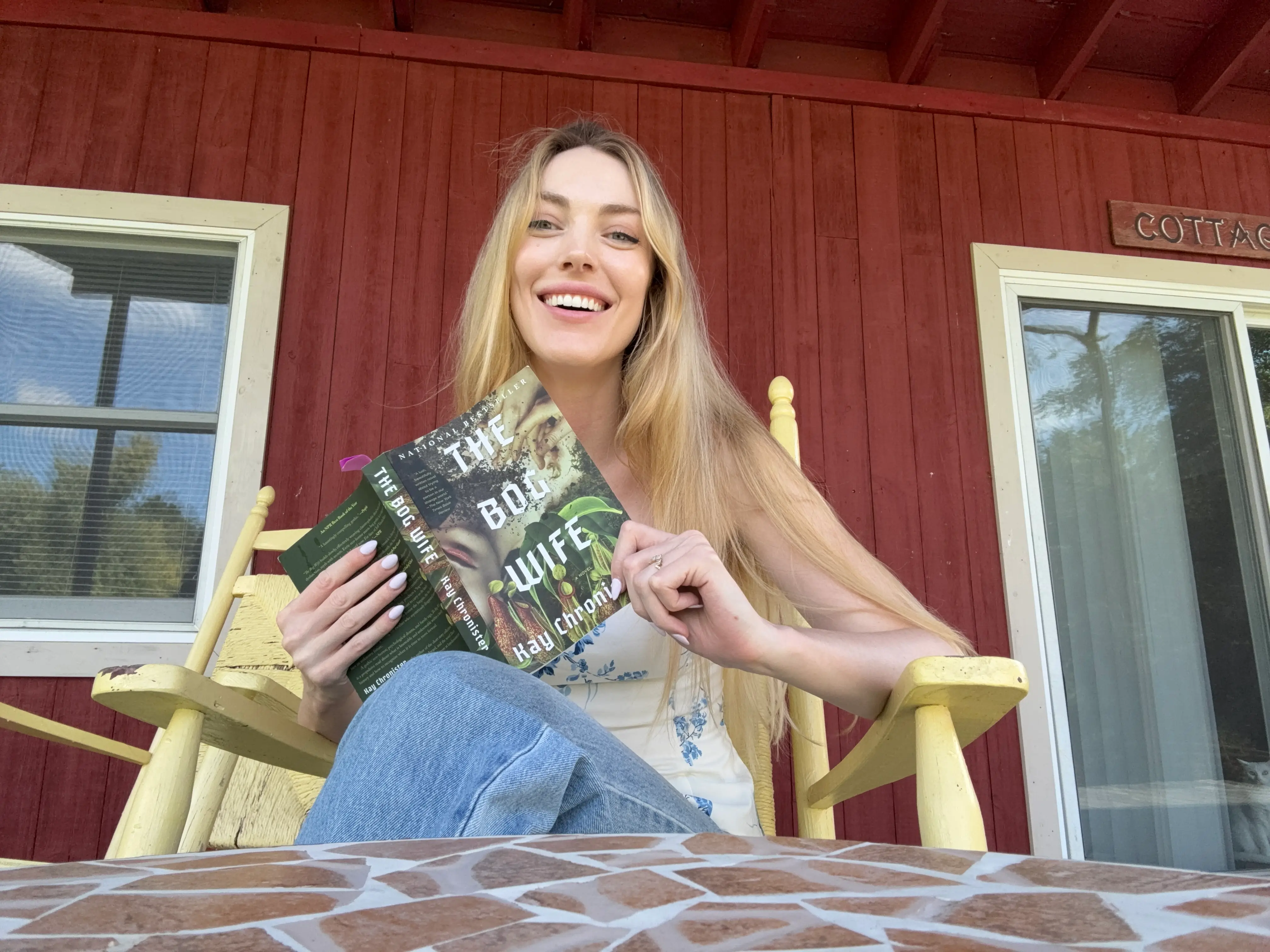 The writer sitting on a porch chair and holding a book, with her cabin visible in the background.