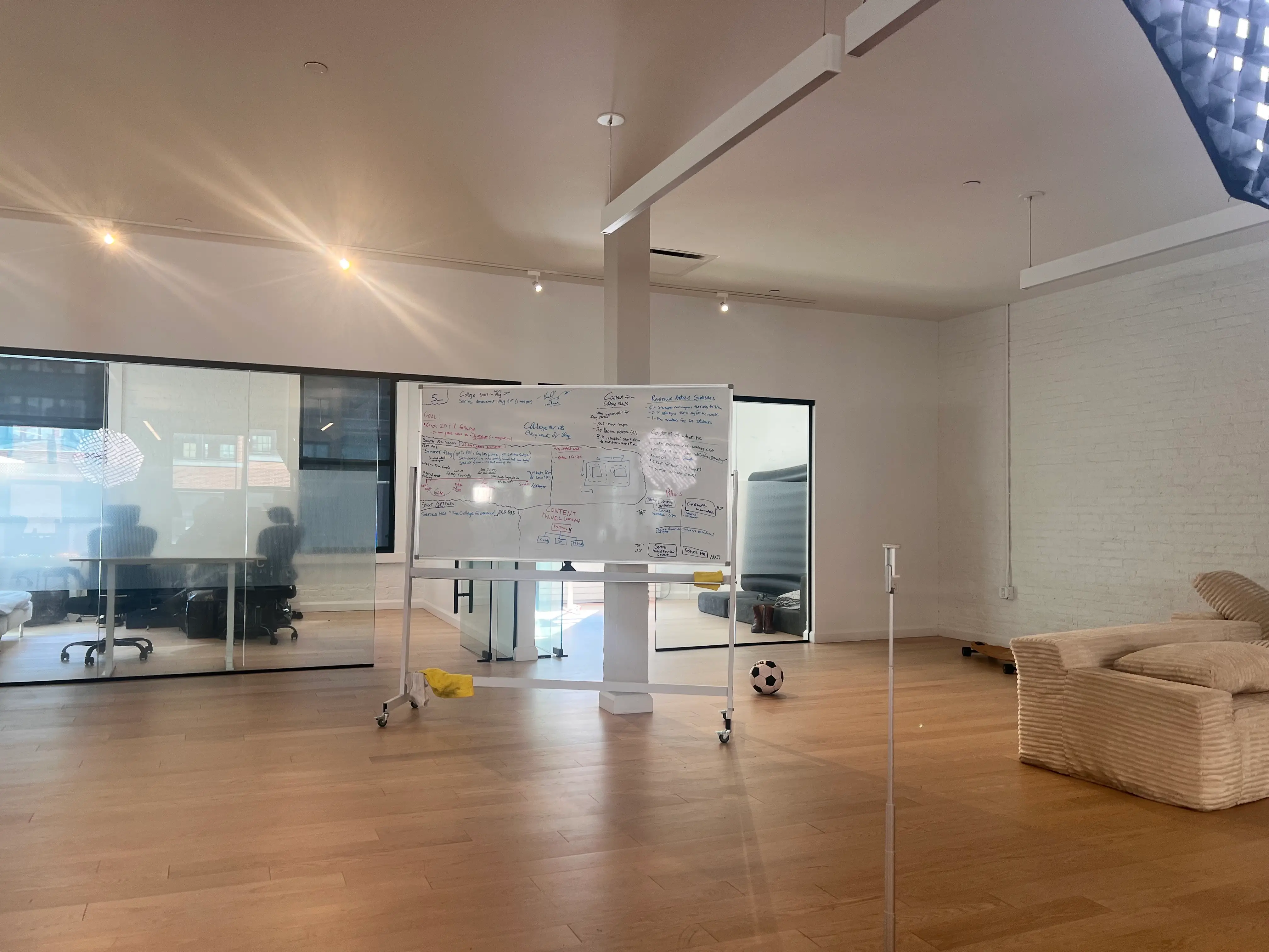 Open office space with glass meeting rooms, a rolling whiteboard, sofa, soccer ball, and wood flooring.