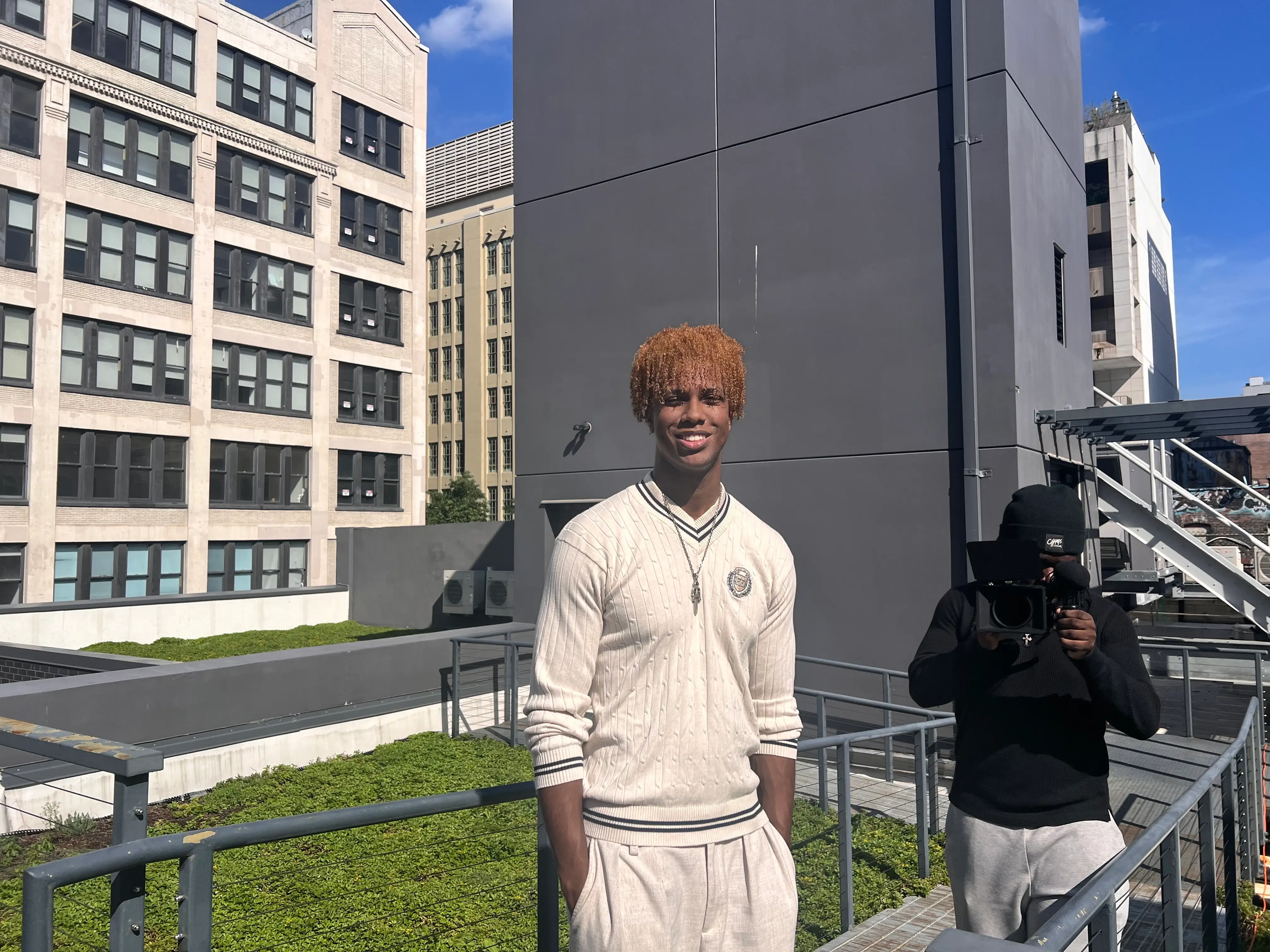 Person in cream sweater stands on a city rooftop while a camera operator films nearby.