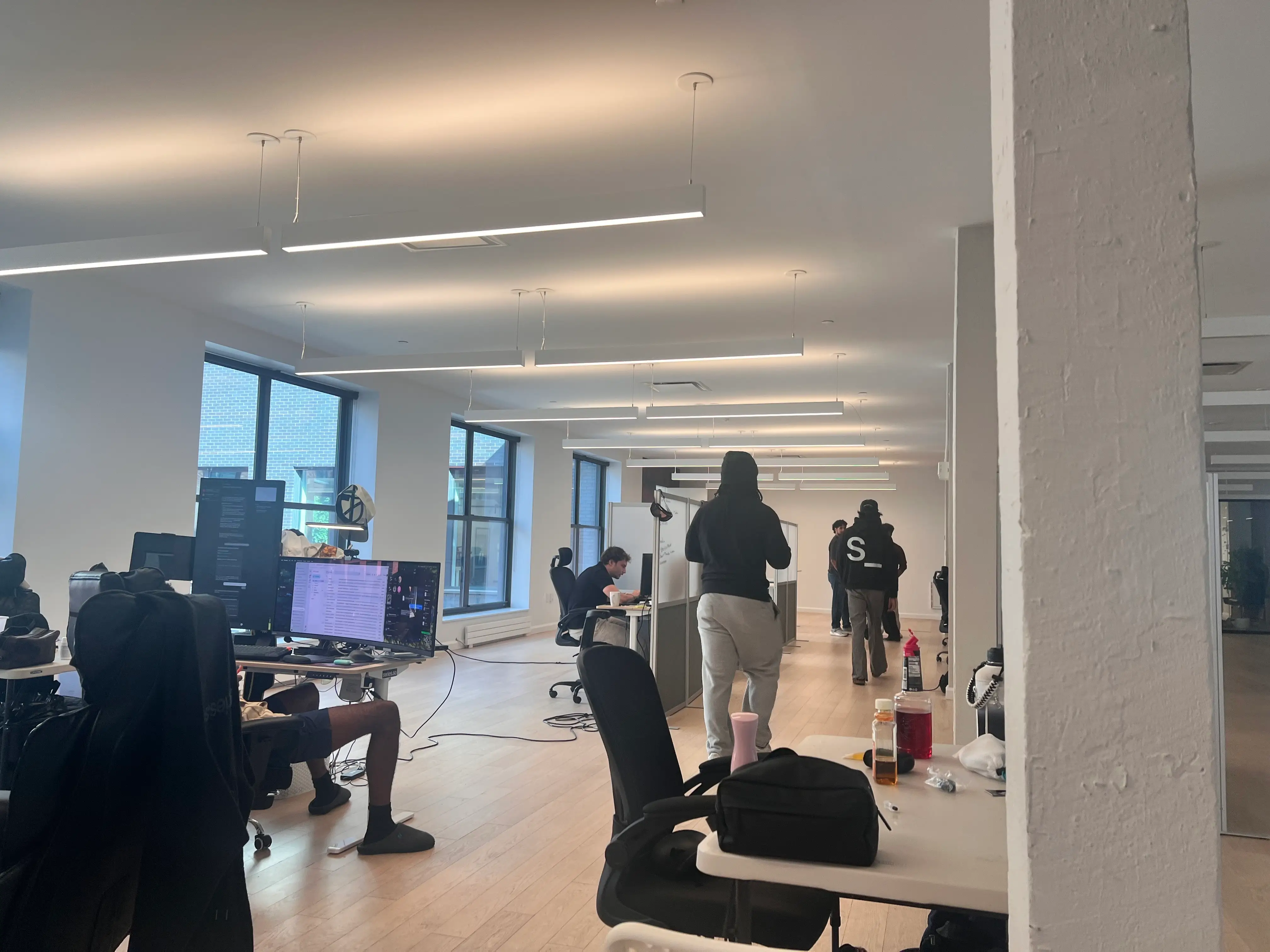Open-plan office with desks, monitors, seated workers, standing crew members, large windows, and linear ceiling lights.