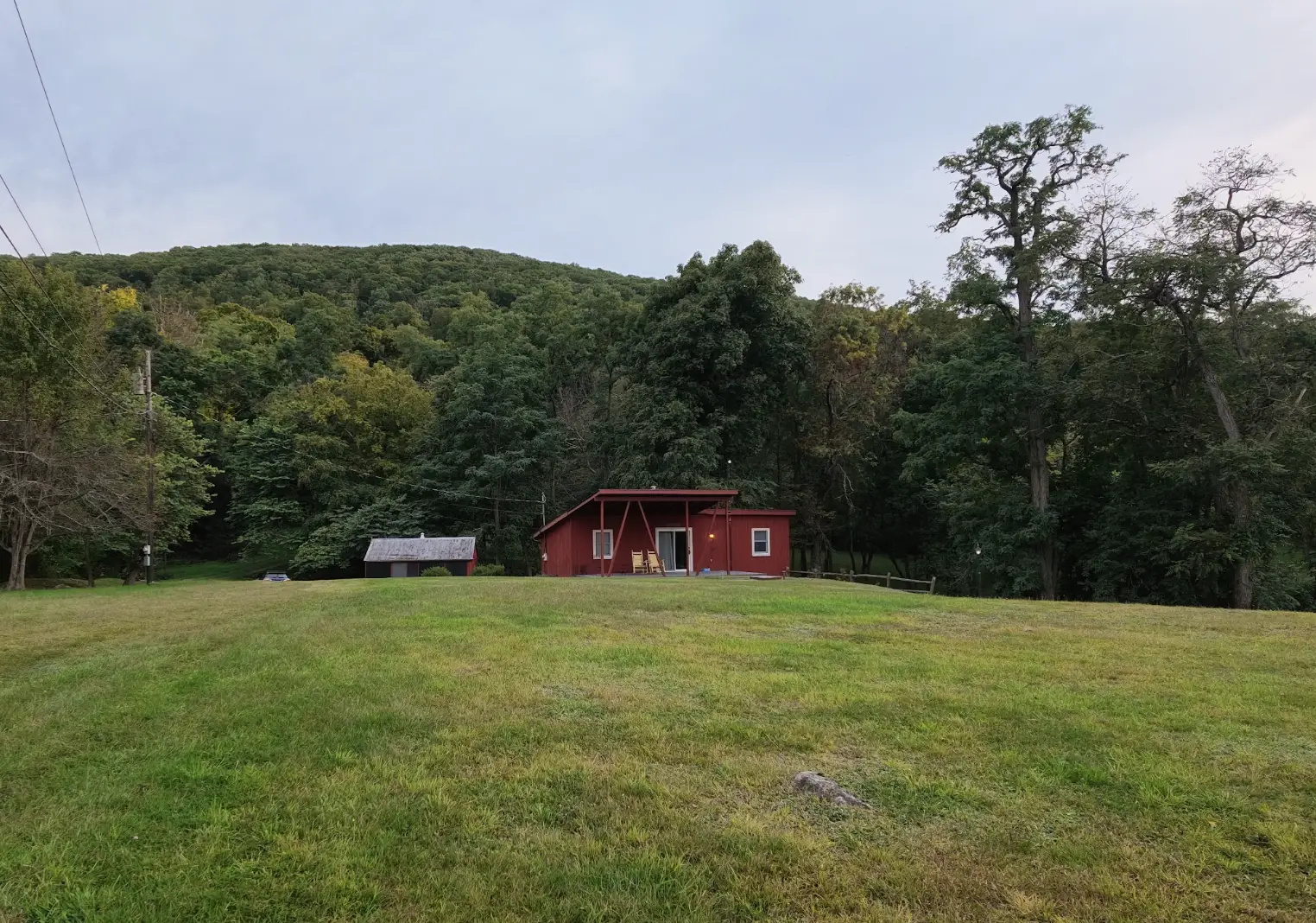 Small red building and shed sit in a grassy clearing below a wooded hillside.