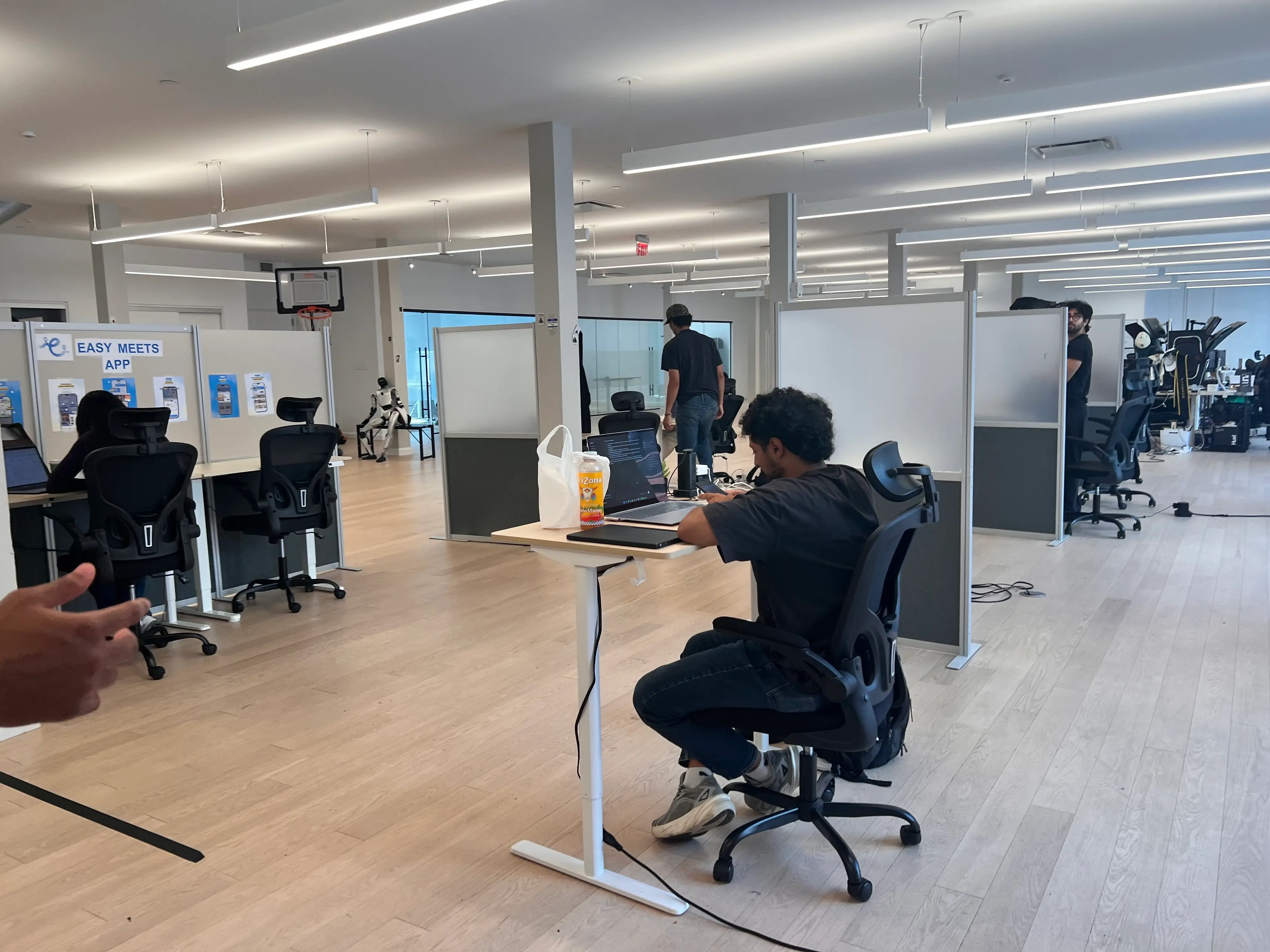 Open-plan office with desks, chairs, partitions, laptops, and several people working under bright ceiling lights.