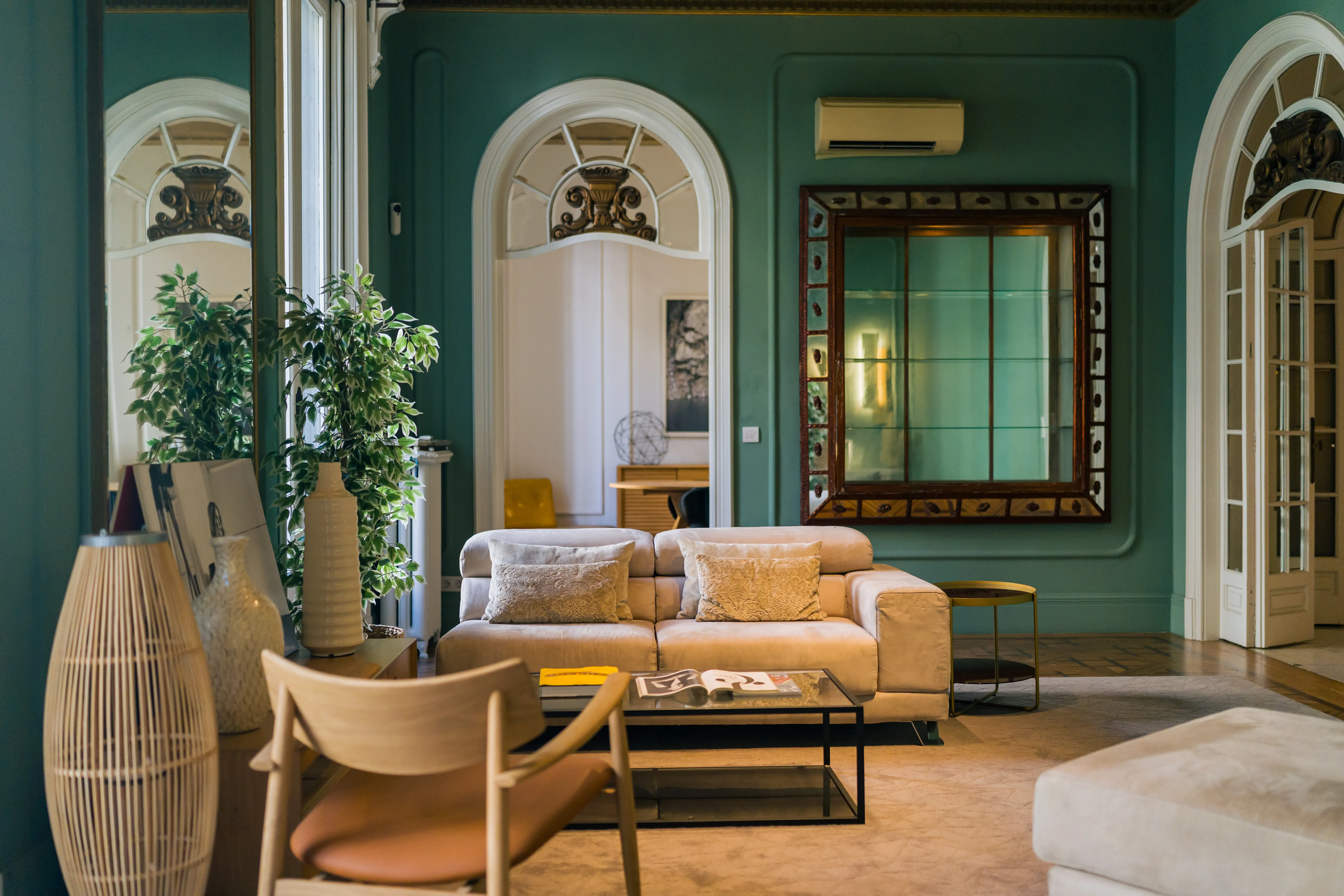 Sophisticated living room in a classic apartment featuring deep green walls, a beige upholstered sofa with plush pillows, an ornate vintage mirror, and arched doorways with decorative moldings.