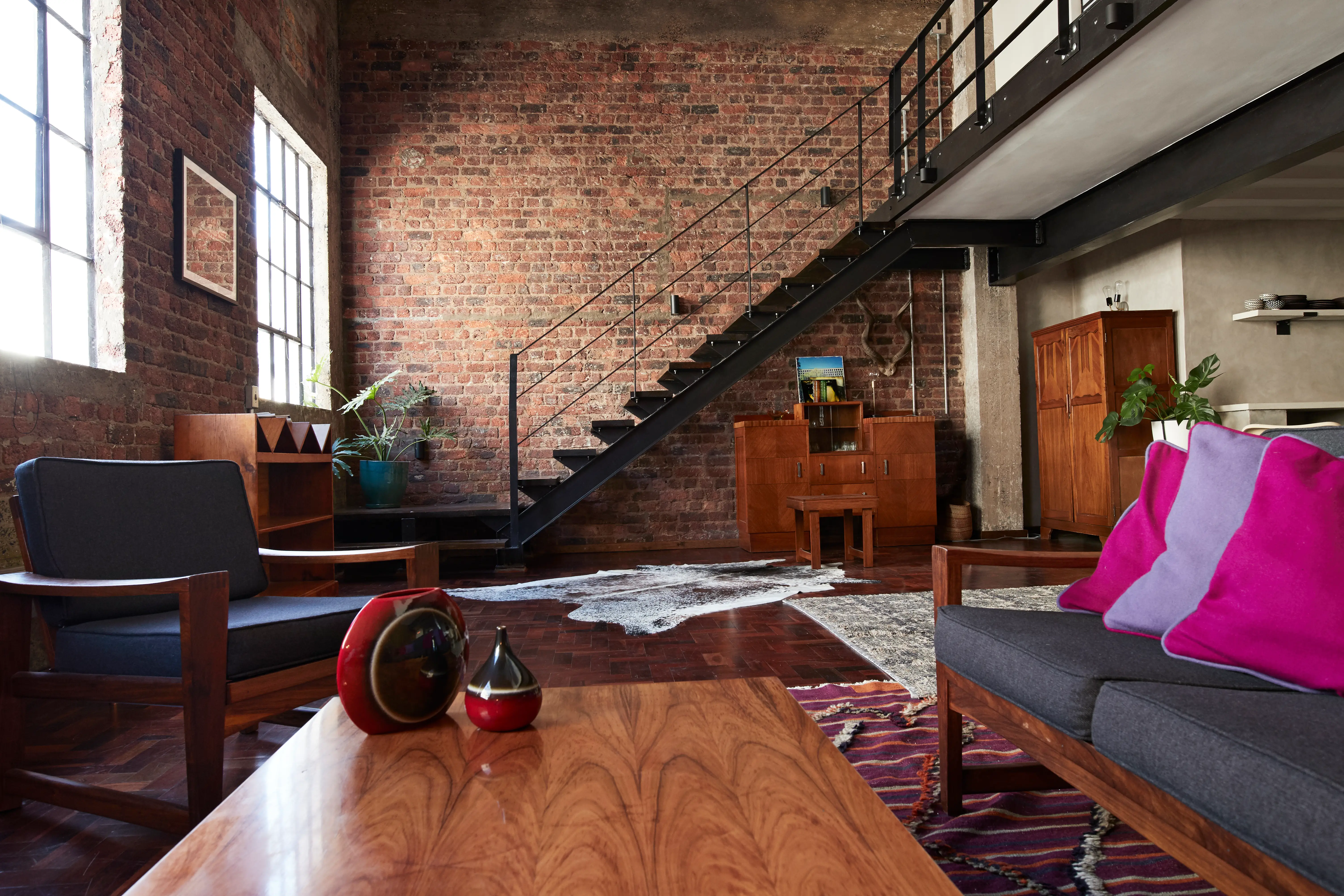 Open living room area with brick walls, wood-frame couch and chair, wood table, large windows