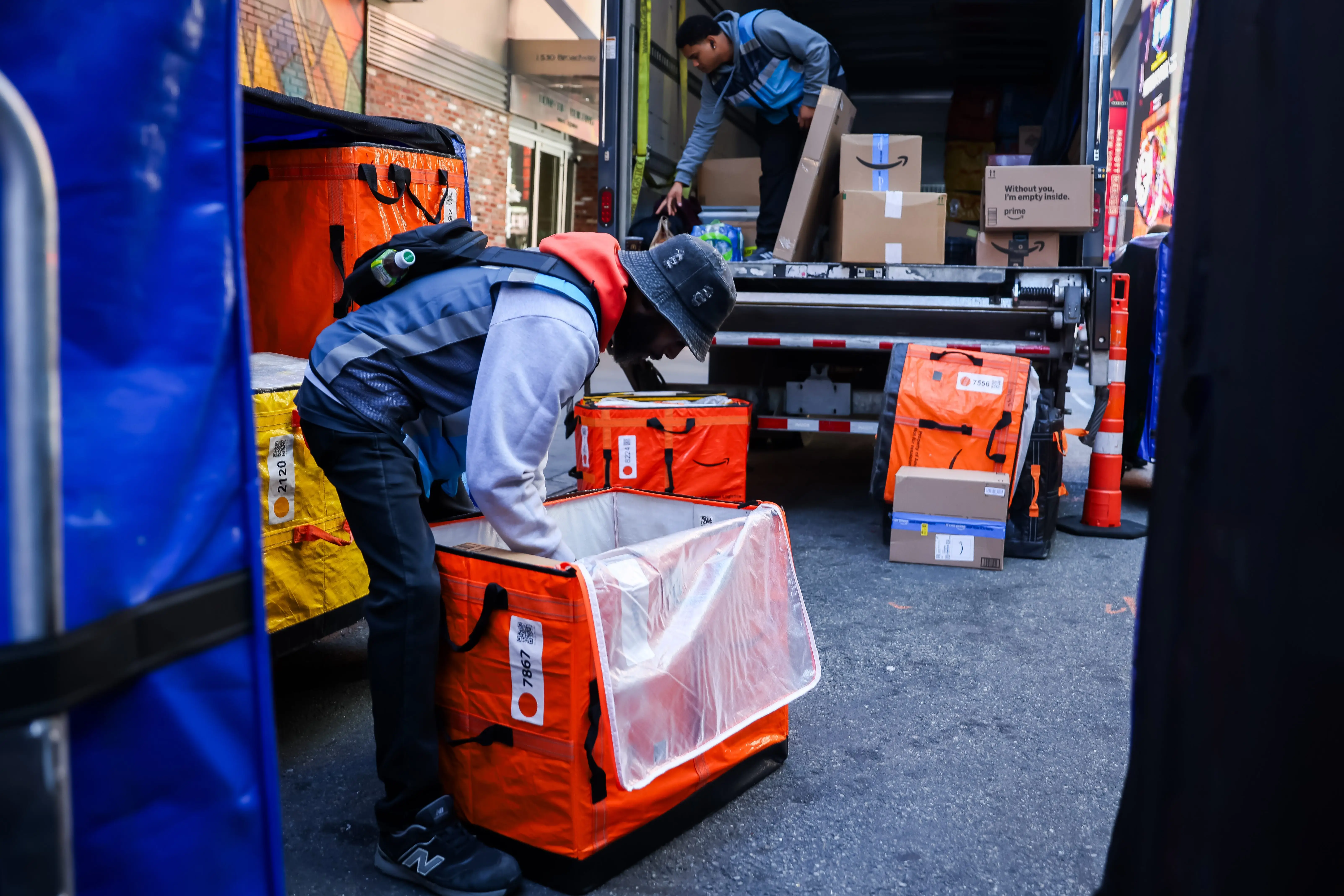 Delivery workers load orange insulated bags and cardboard boxes into the back of a truck on a city street.