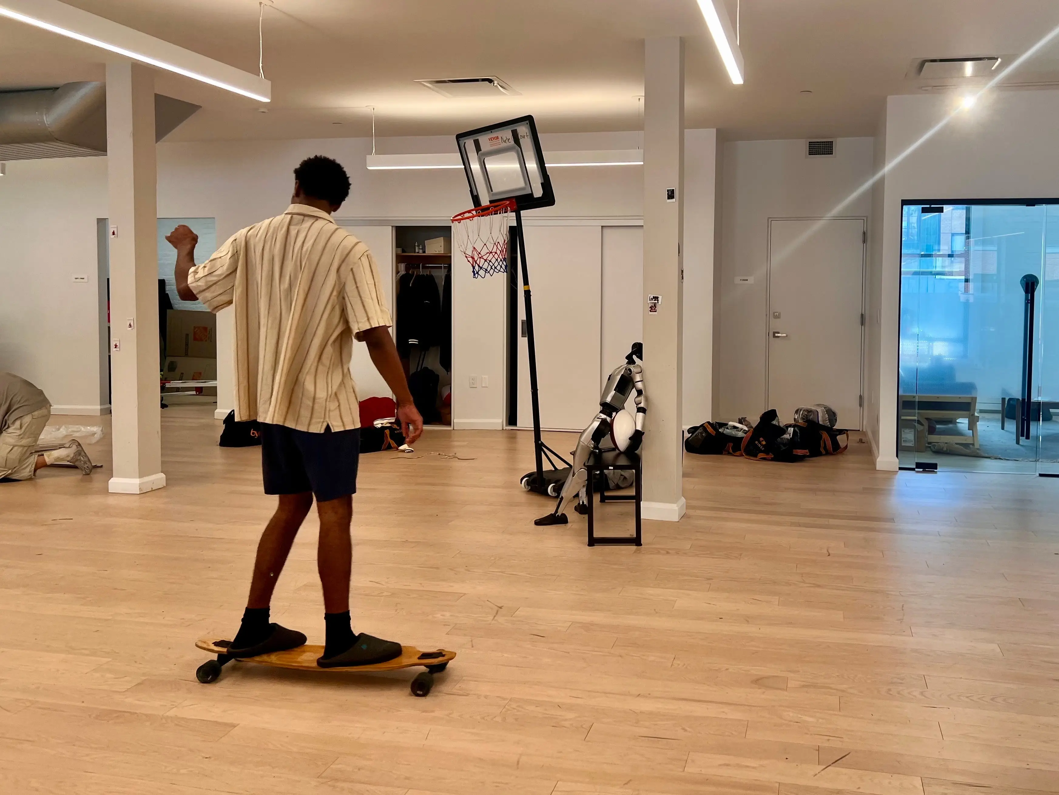 Person on a skateboard inside an open room with a portable basketball hoop, robot, and glass-walled office area.