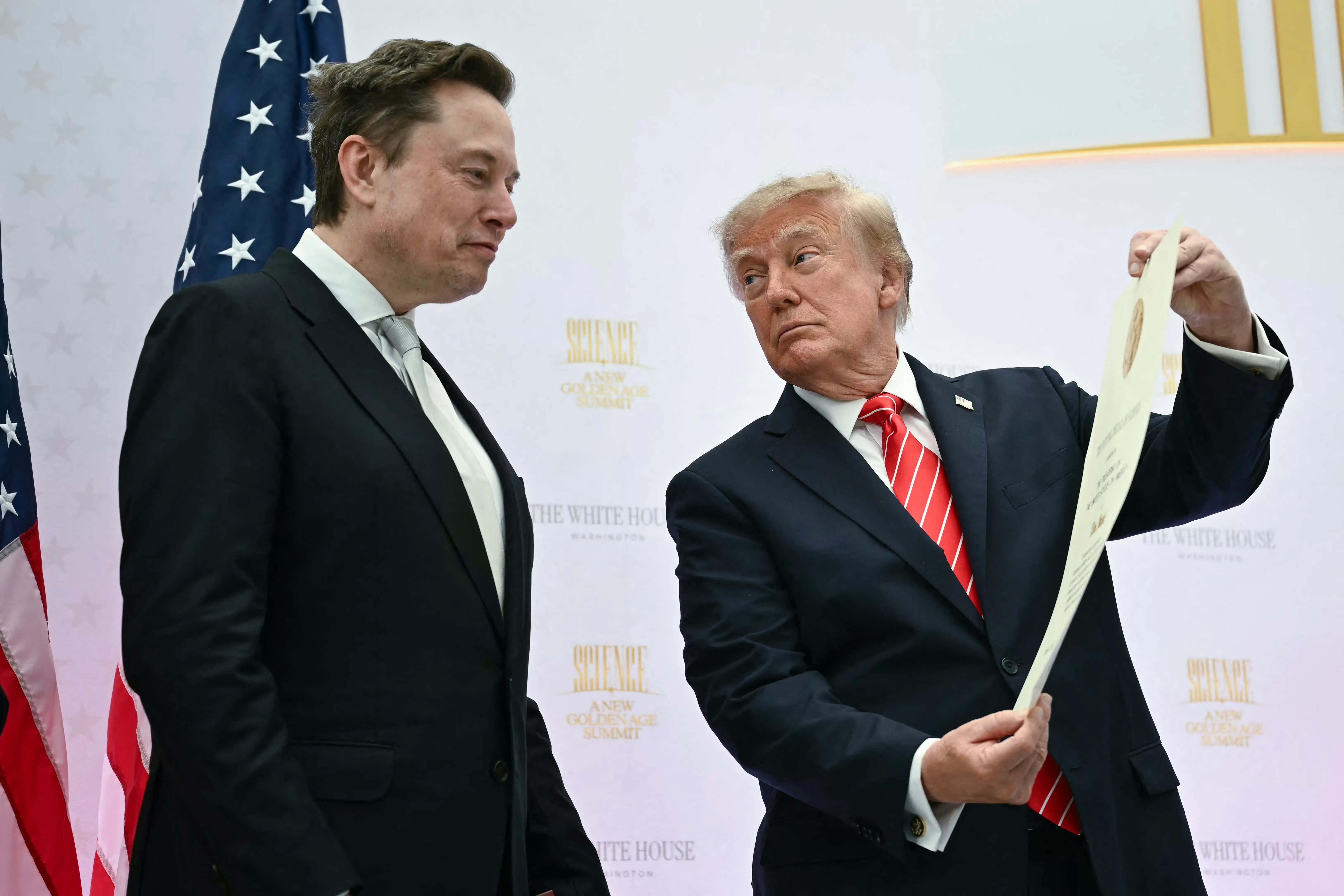 Donald Trump holds a document beside Elon Musk in front of a White House backdrop and U.S. flag.
