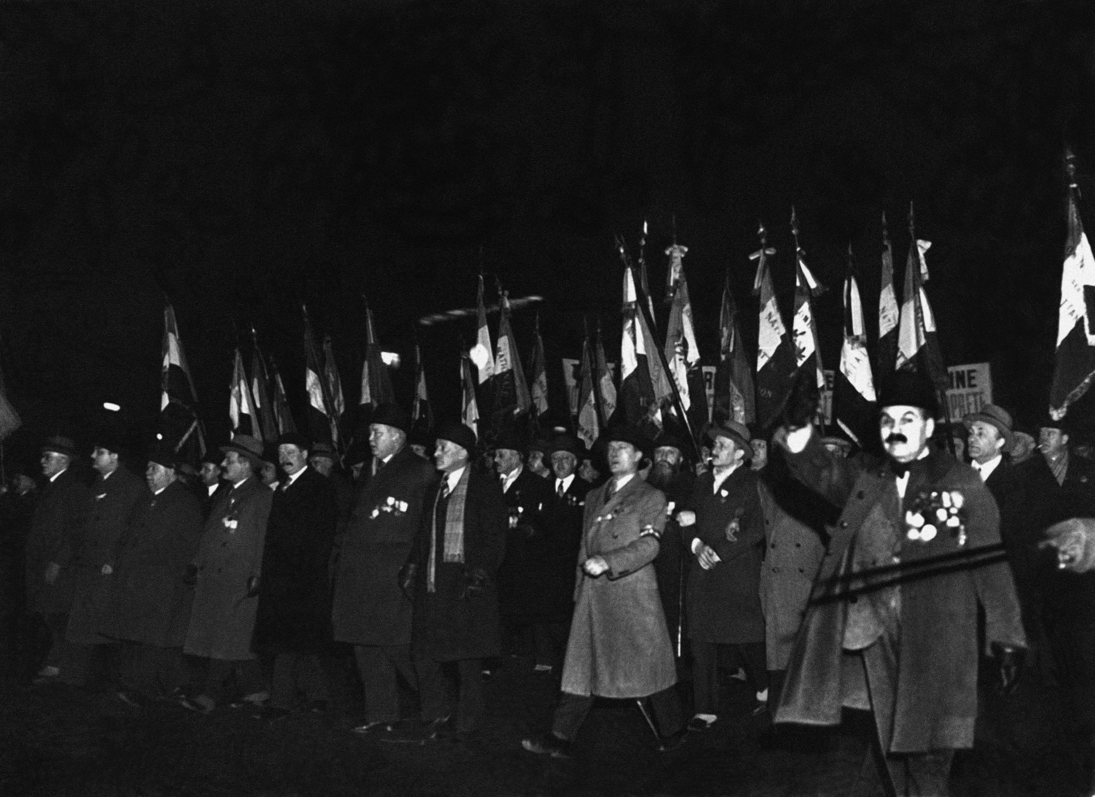 Nighttime black-and-white scene of marchers carrying many flags, with several people wearing medals and heavy coats.