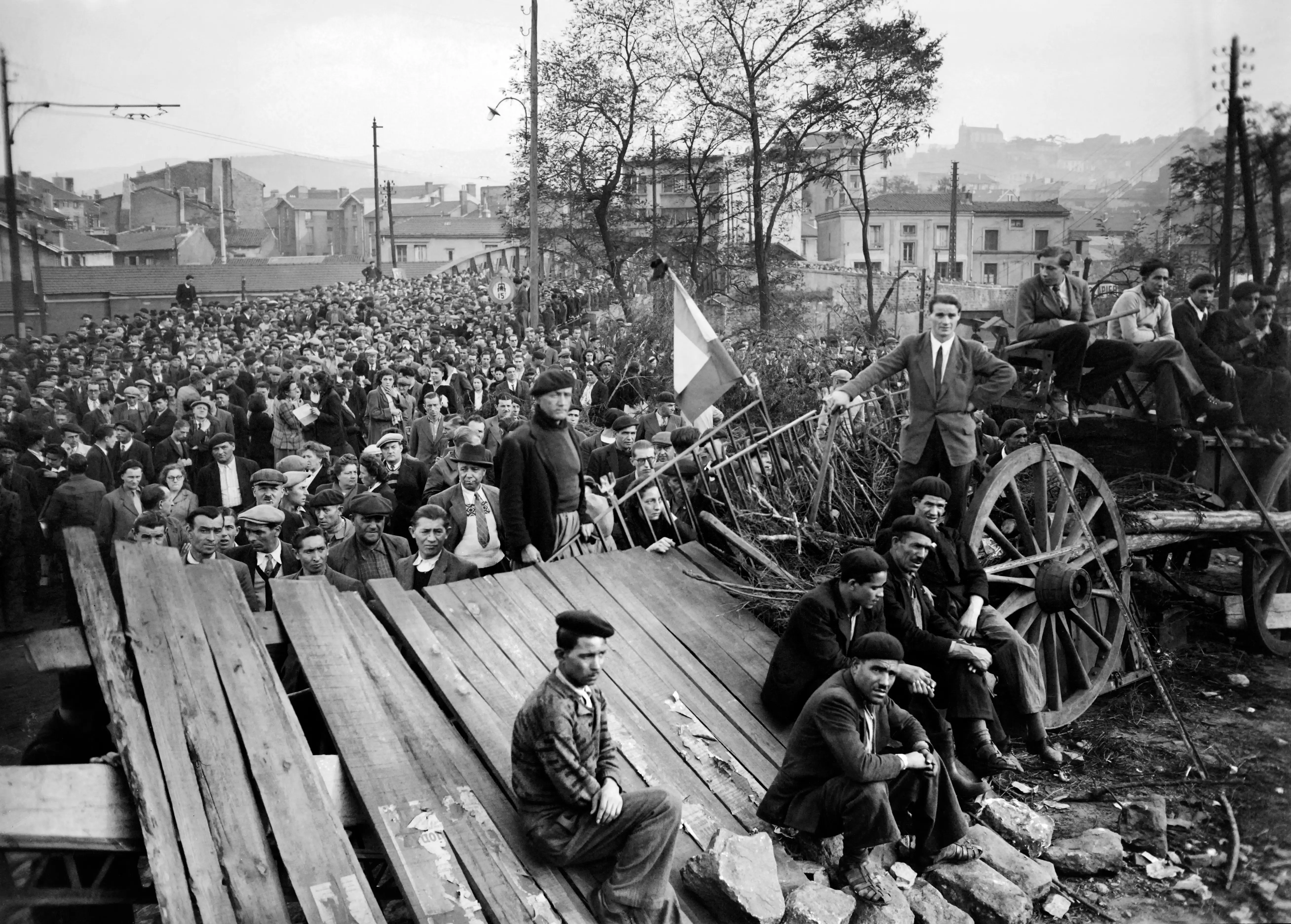 Large crowd gathers near wooden planks and a horse-drawn cart in a black-and-white urban street scene.