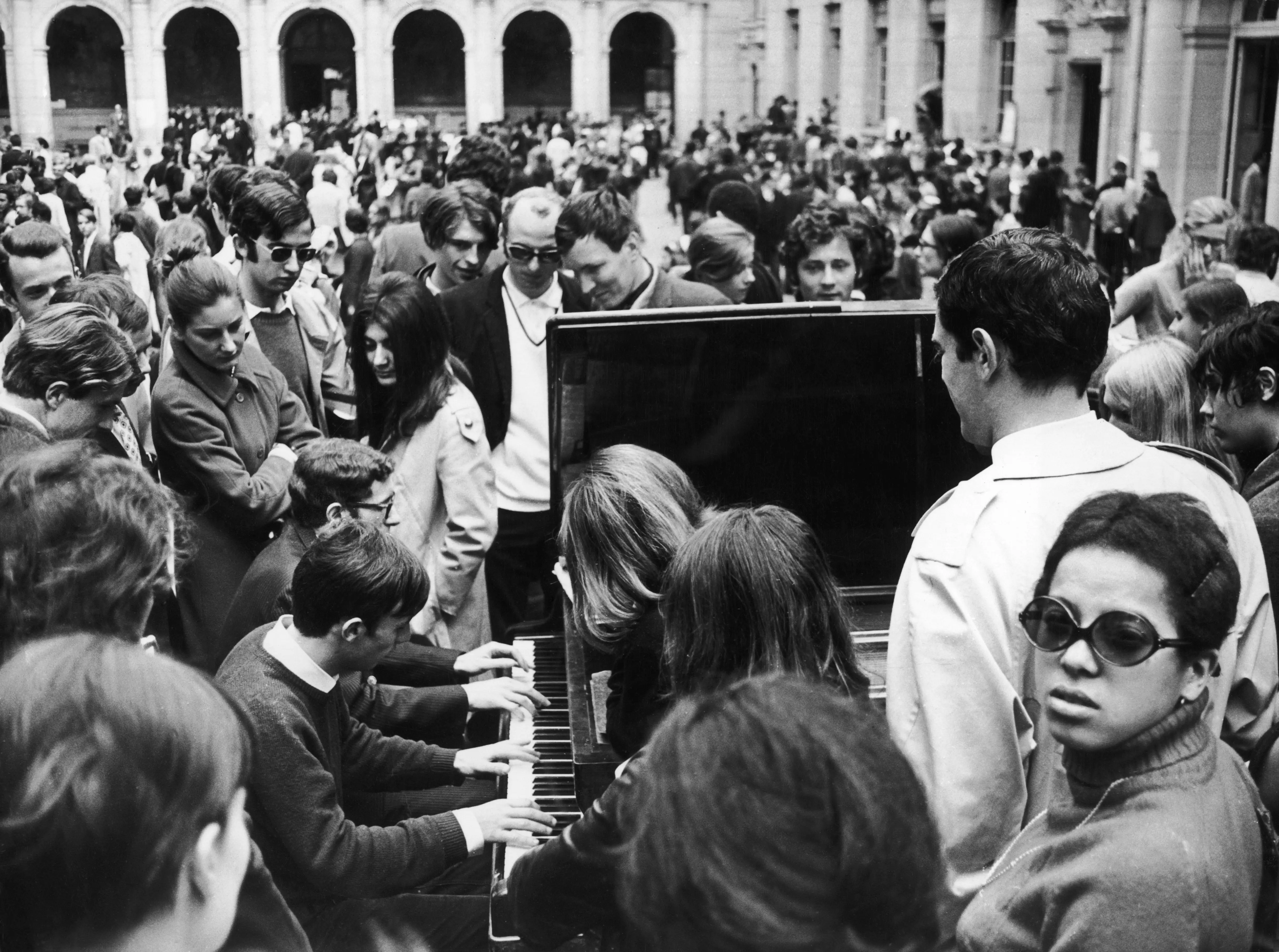 Crowd gathers around a pianist playing an upright piano in a busy arched courtyard.
