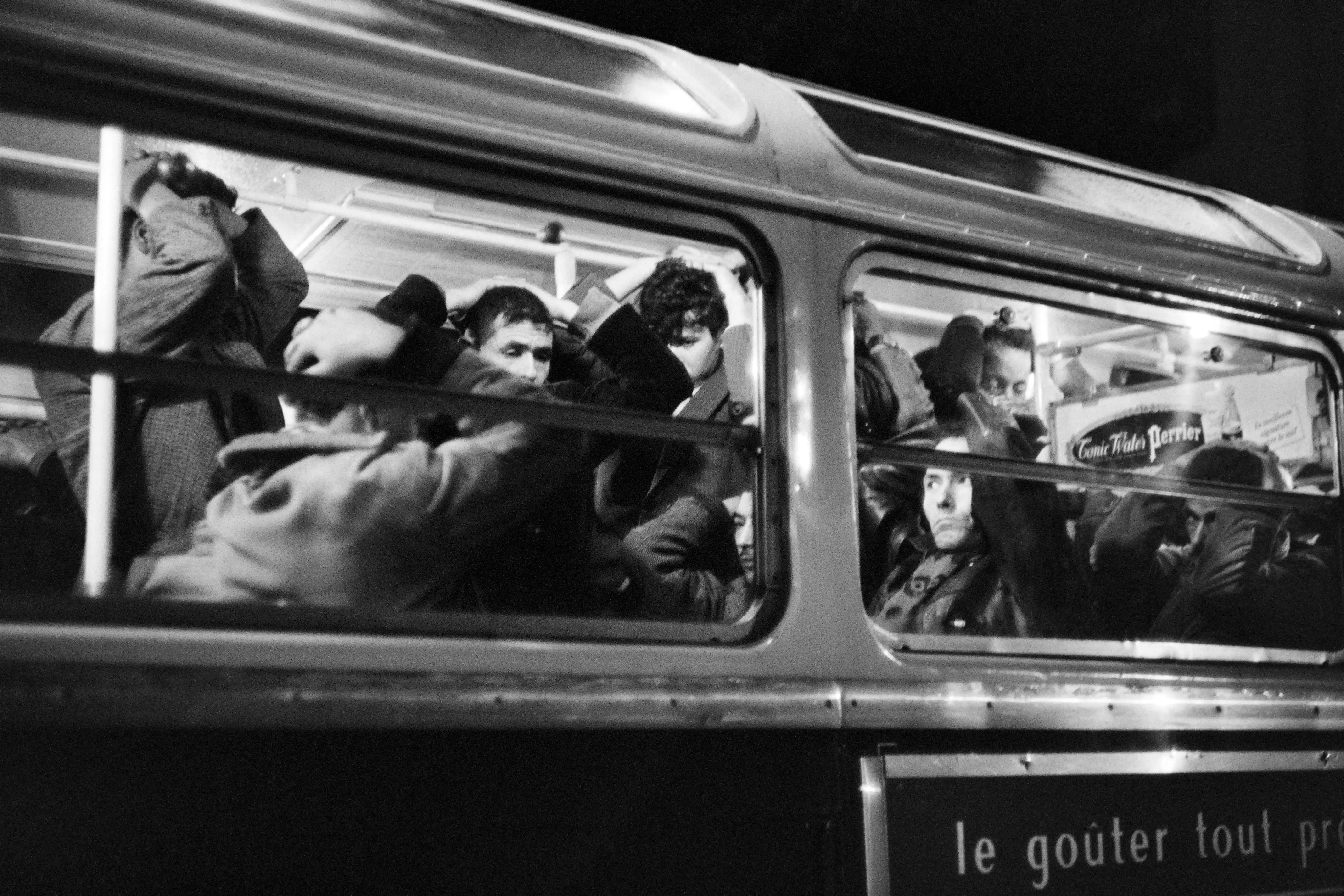 Passengers crowd inside a city bus at night, seen through large windows in a black-and-white street scene.