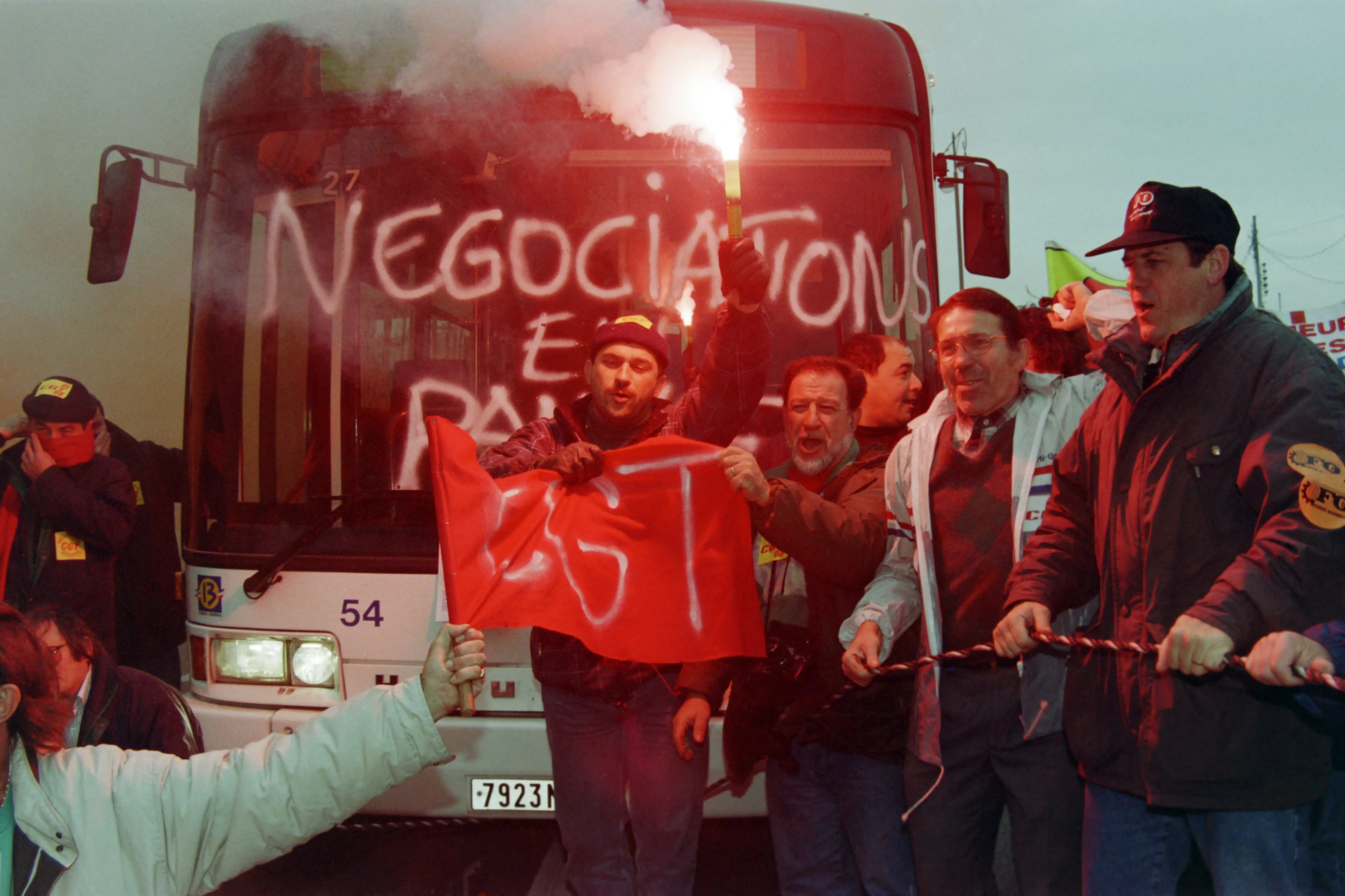 Demonstrators holding a red flag and flare stand in front of a bus marked with white graffiti.