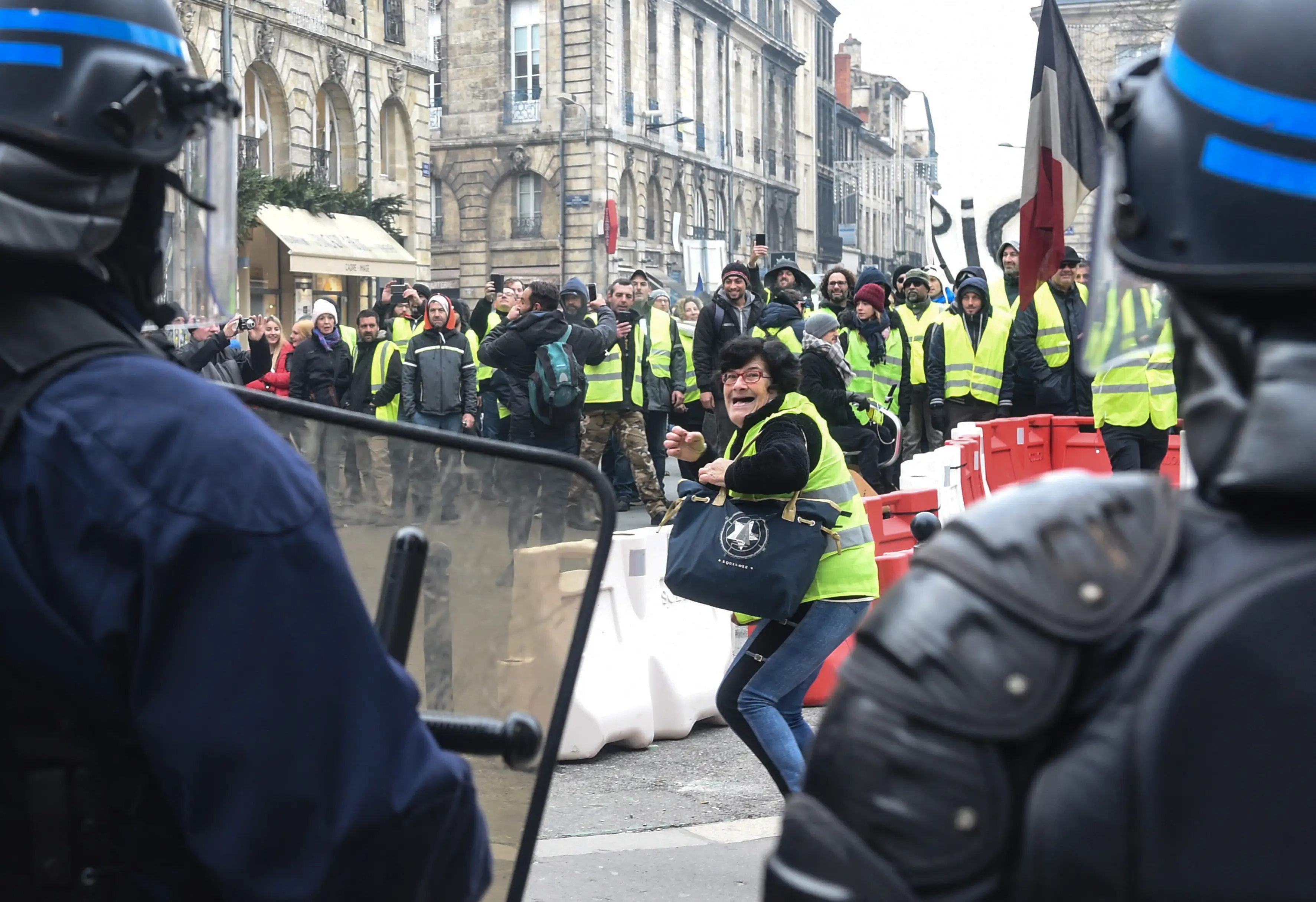 Yellow-vest protesters face riot police behind street barricades during an urban demonstration with a French flag visible.