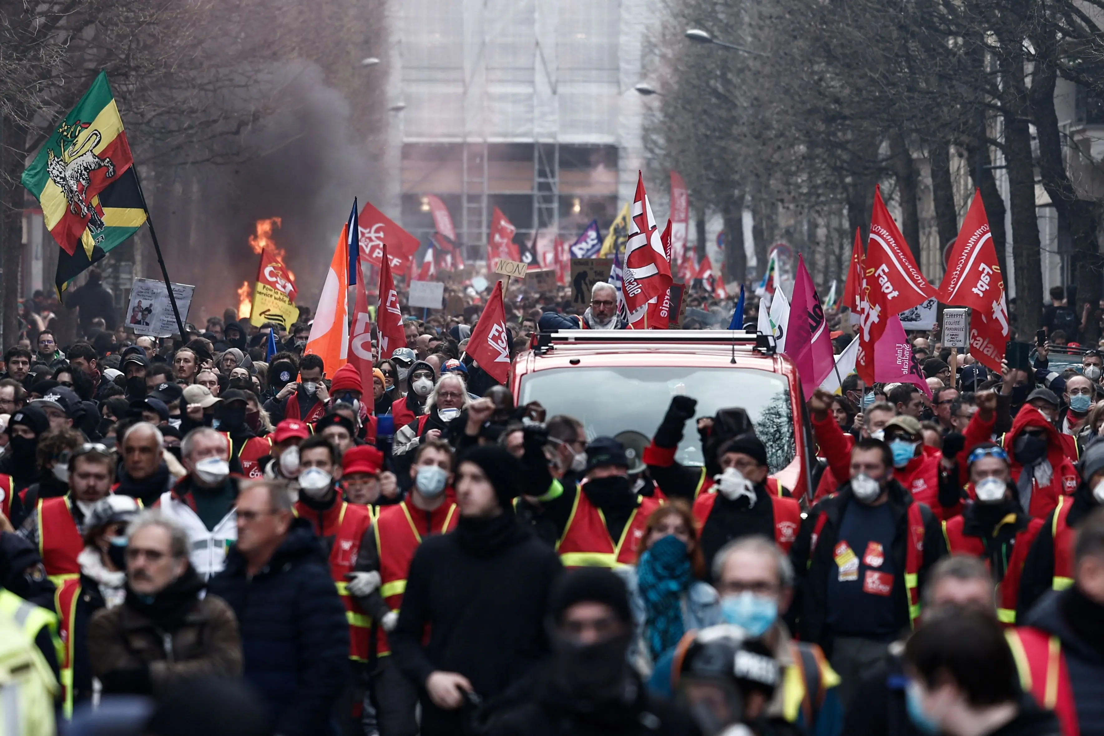 Crowd of demonstrators with CGT flags and yellow vests marches through a smoky city street.