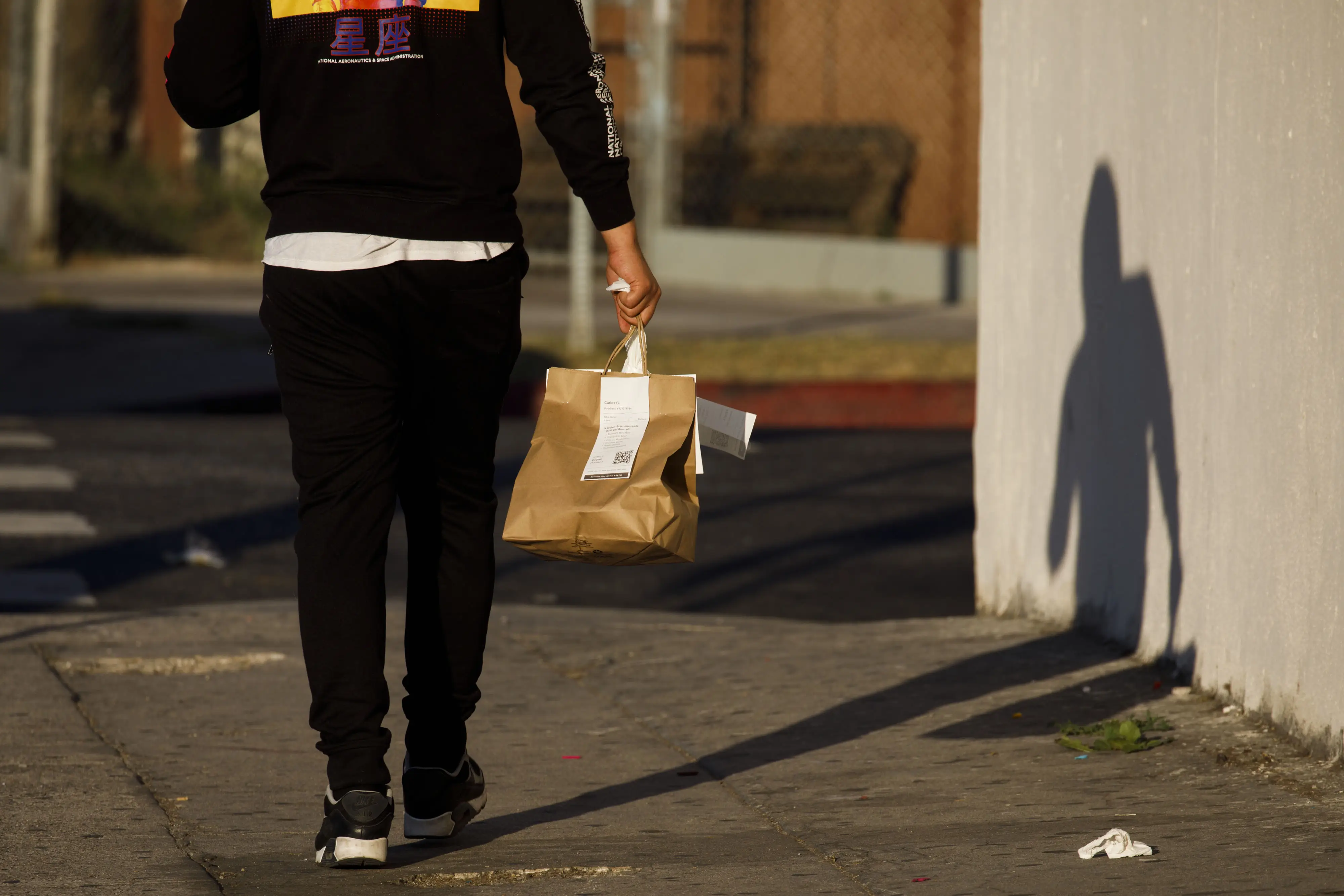 Person walking on a sunlit sidewalk carrying a brown paper takeout bag beside a wall casting a long shadow.
