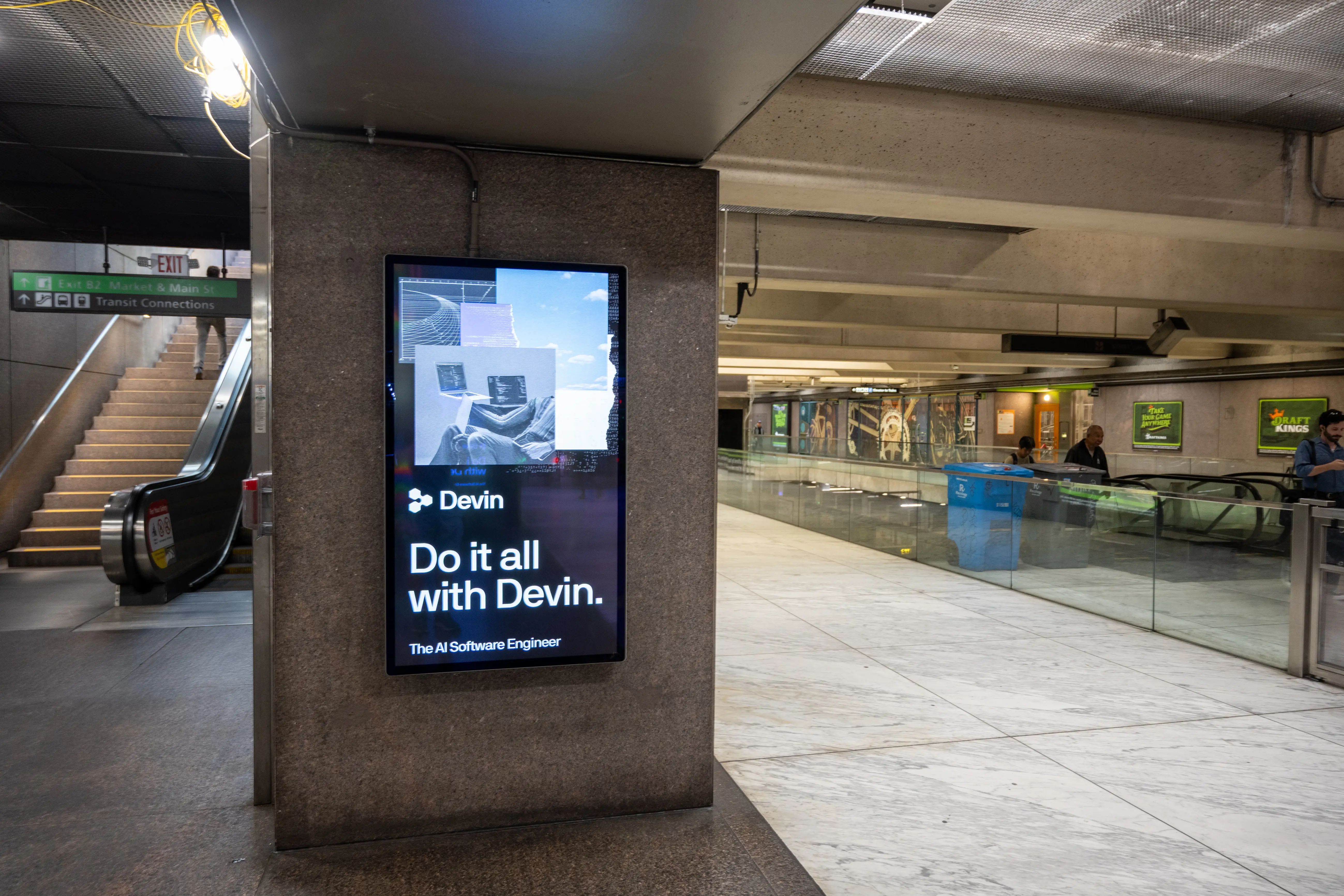 Digital advertisement for Cognition's Devin AI software engineer beside escalators near Exit B2 on the concourse at Embarcadero BART and Muni Metro station, San Francisco, California, August 27, 2026.