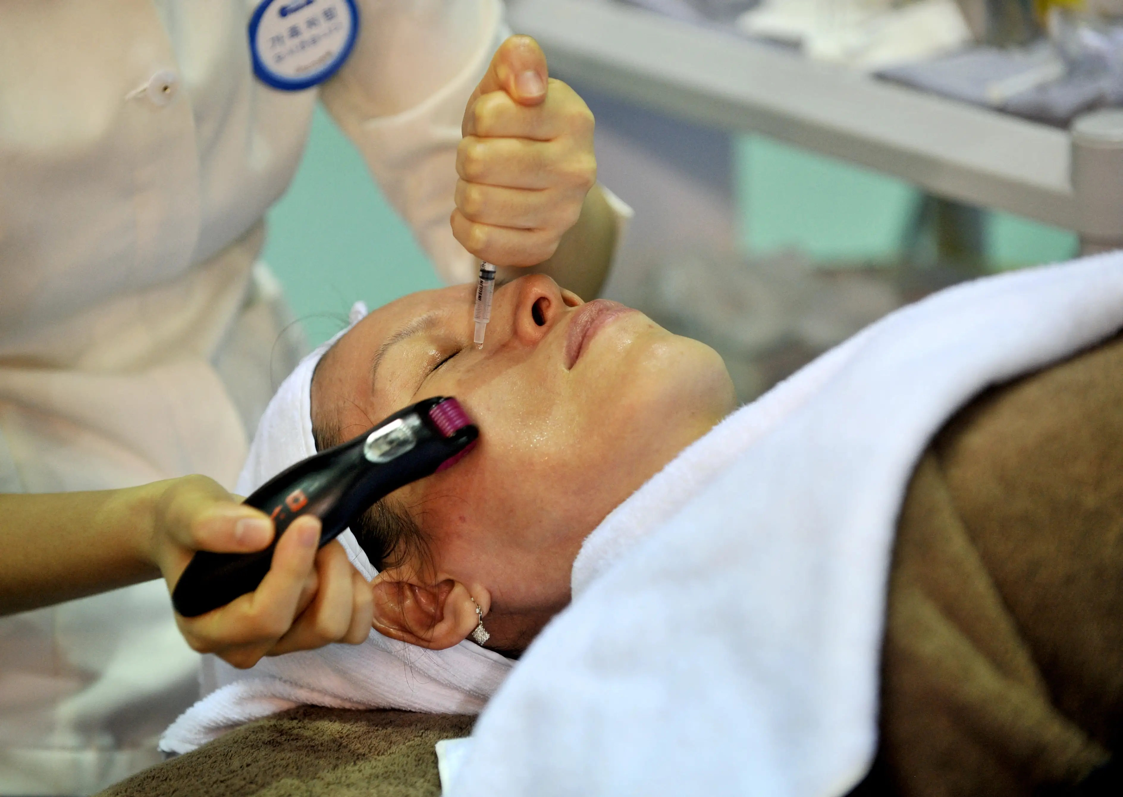 A client lies under towels while a practitioner uses a syringe and handheld device during a facial treatment.