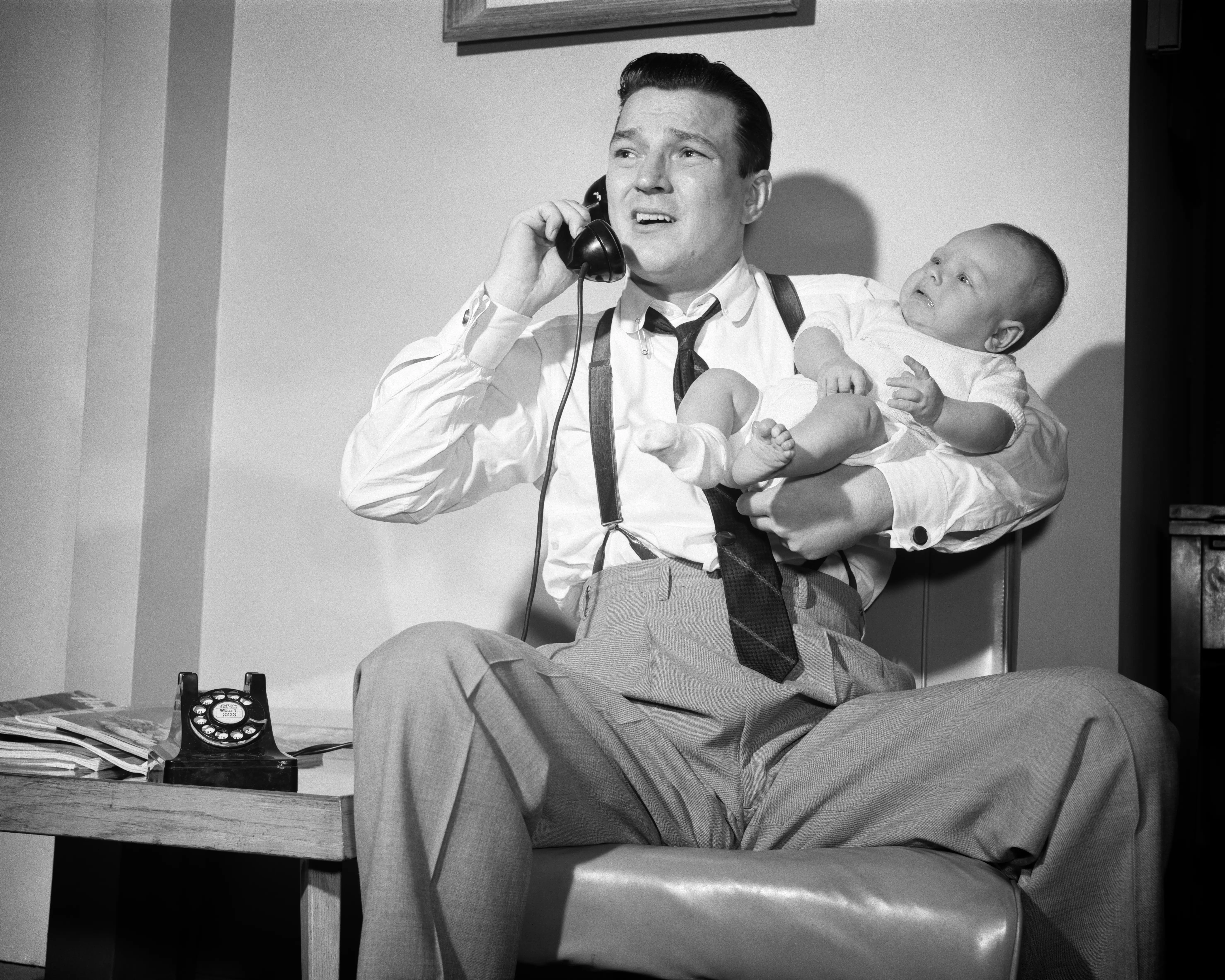 1950s distressed father holding baby while talking on telephone. (Photo by Debrocke/ClassicStock/Getty Images)