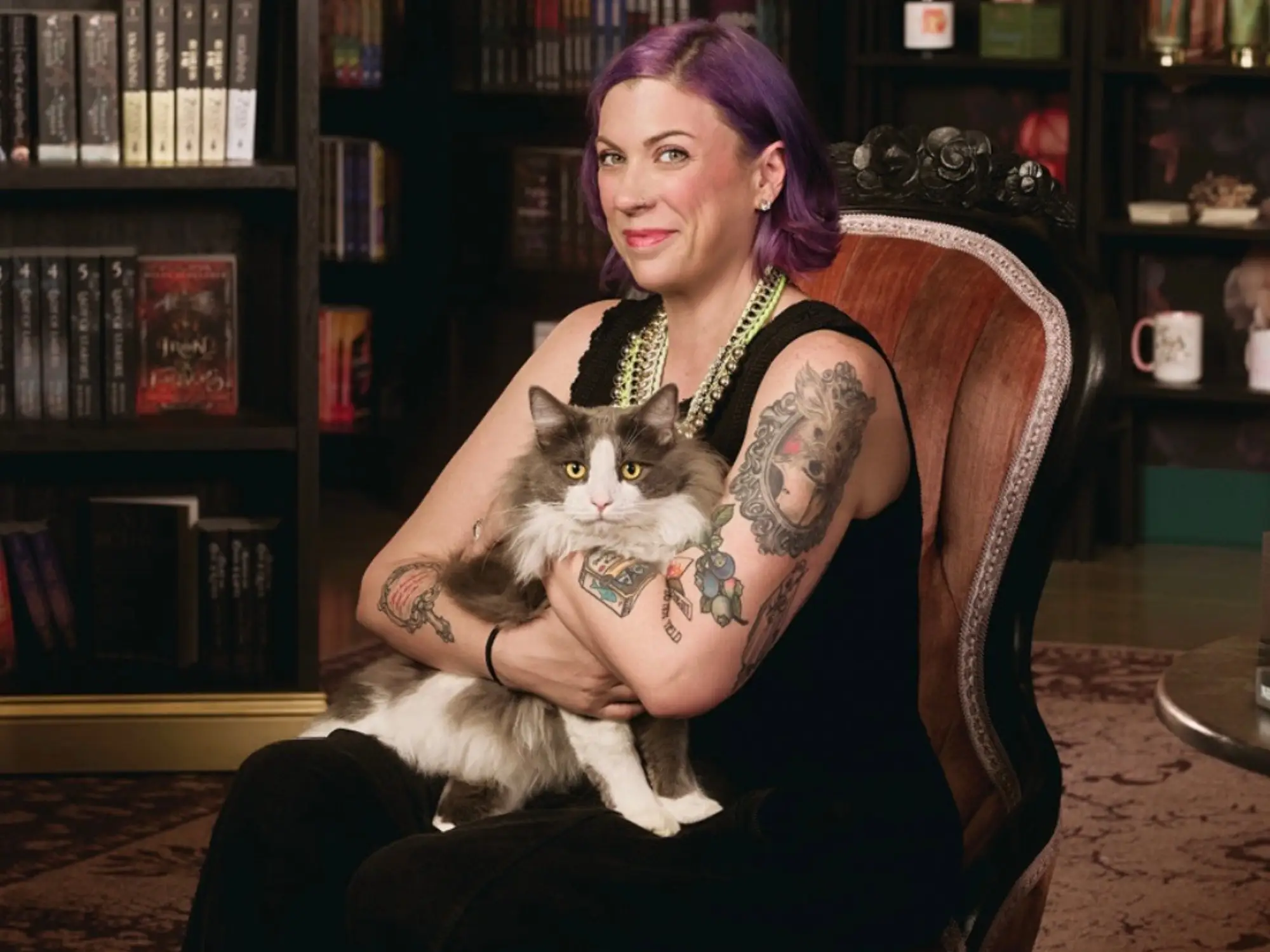 Kate McNeil is sitting in a reading chair, holding a cat in her lap, with a bookshelf in the background.