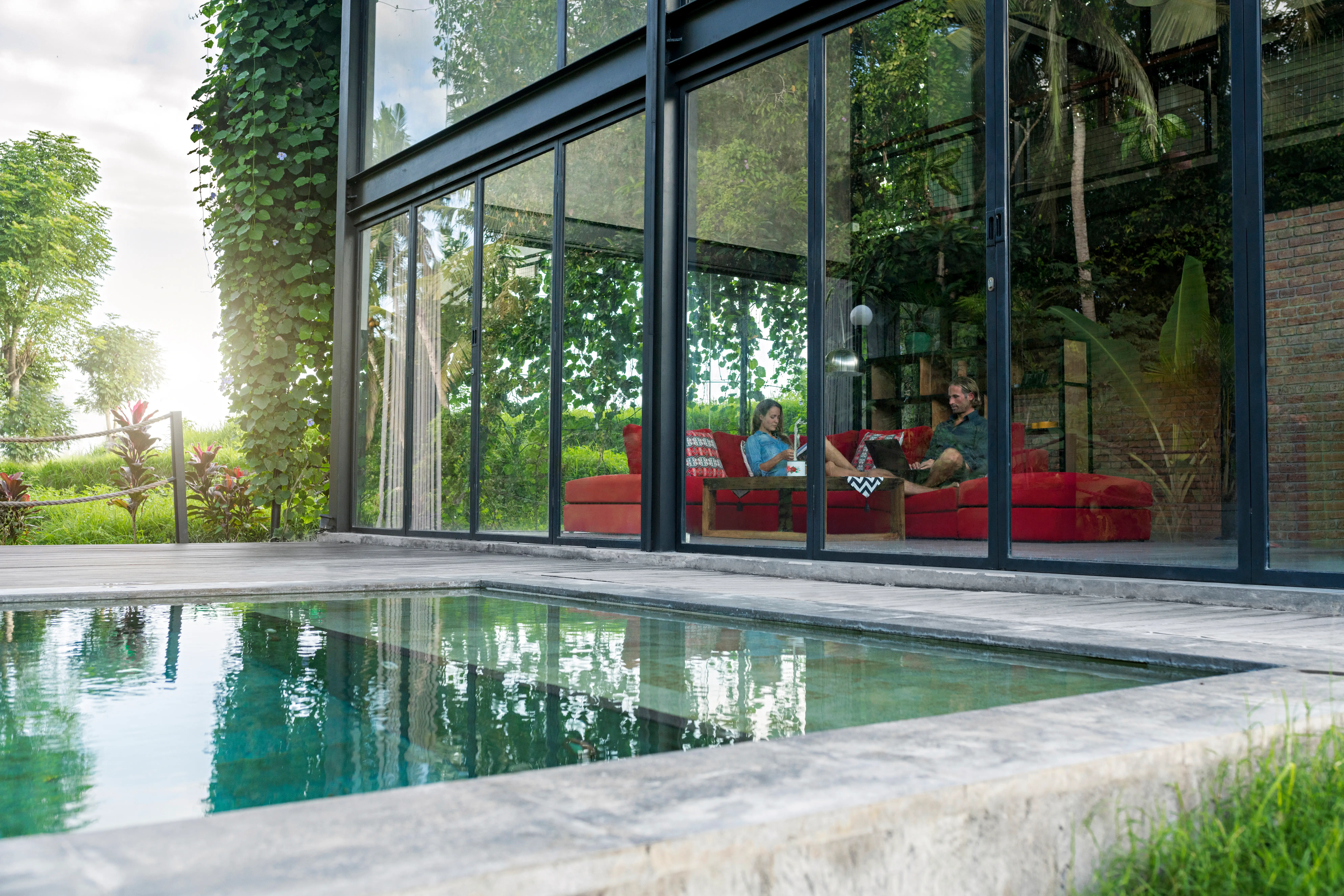 Two people sit on red sofas inside a glass-walled modern home. A pool sits beside it, surrounded by trees.