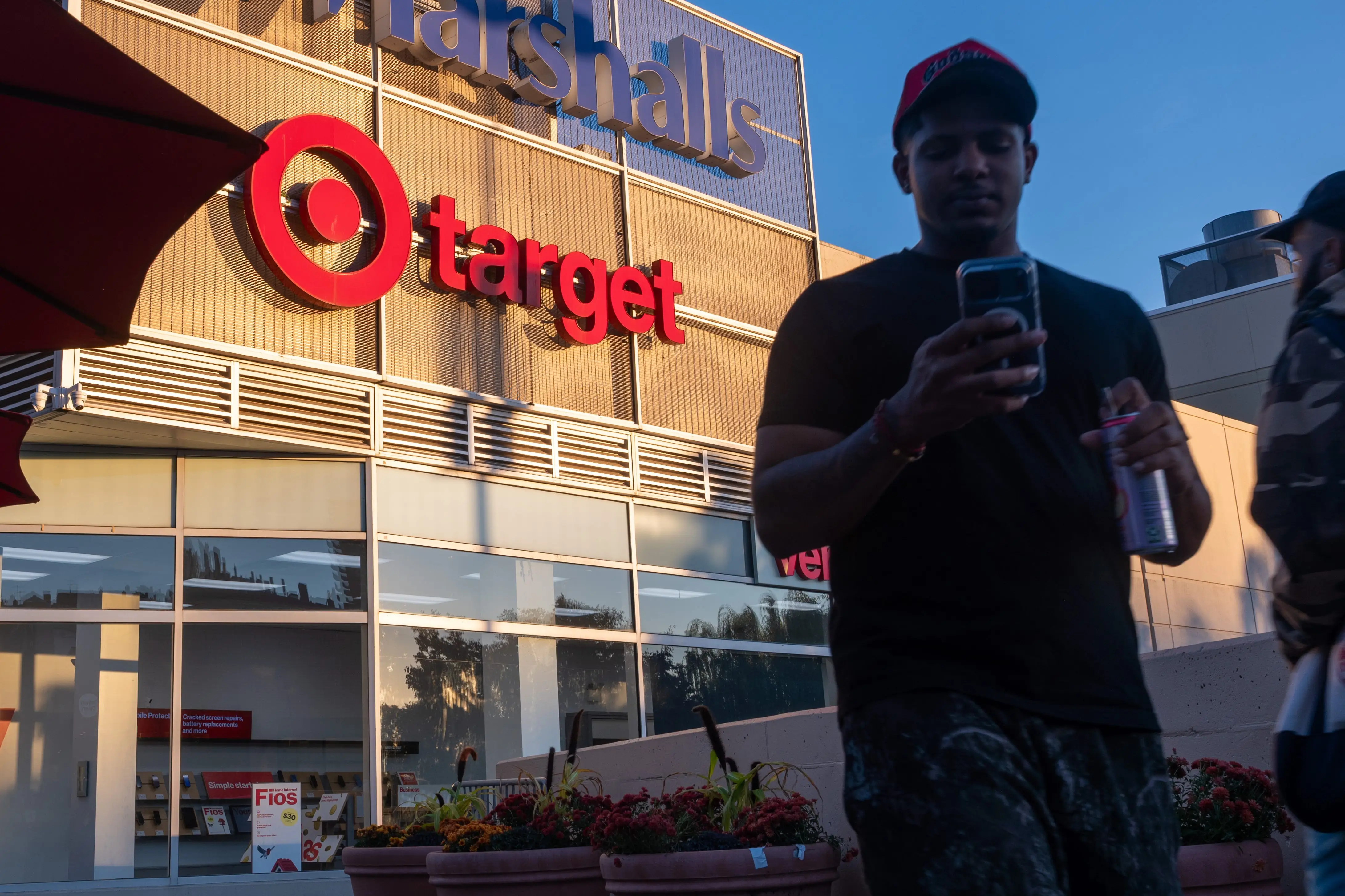Target and Marshalls storefronts are seen at dusk with pedestrians, a red umbrella, and planters in the foreground.