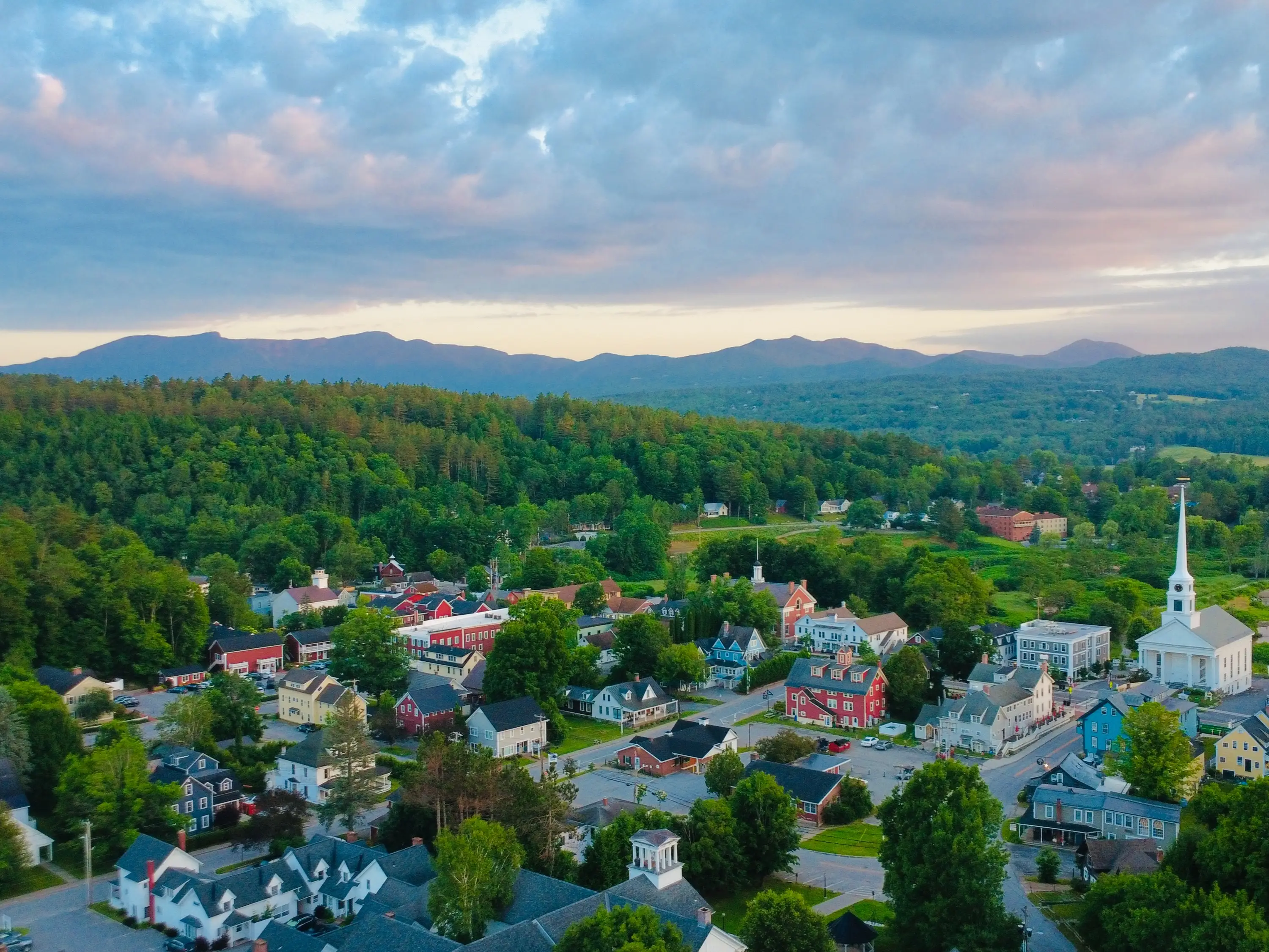 Aerial view of a small town with a white church steeple, forested hills, and mountains under a cloudy sky.