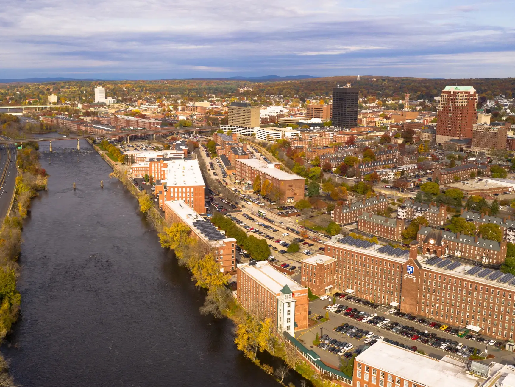Aerial view of a riverfront city with brick mill buildings, downtown towers, highways, and autumn trees.