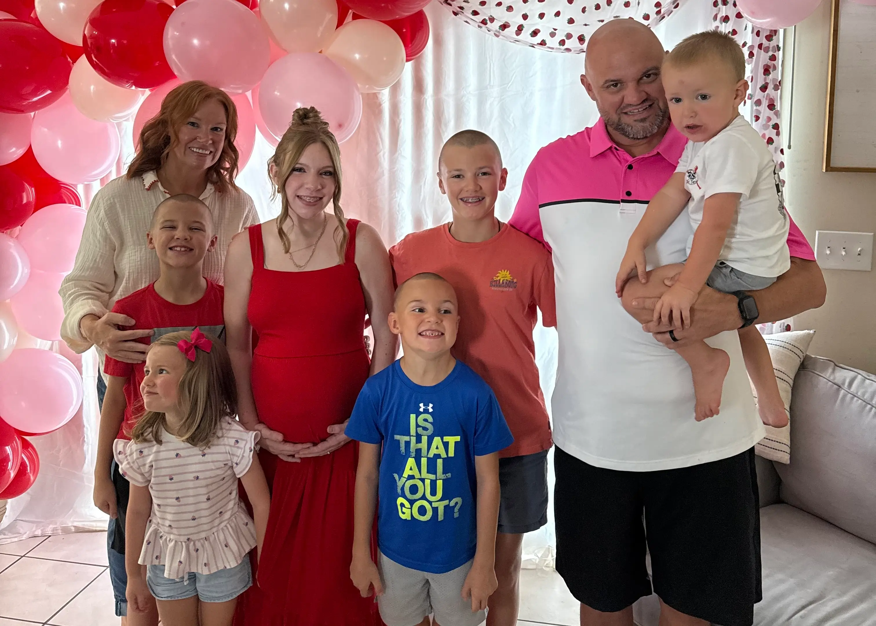 Group of eight people posing indoors in front of red and pink balloon decorations at a celebration.
