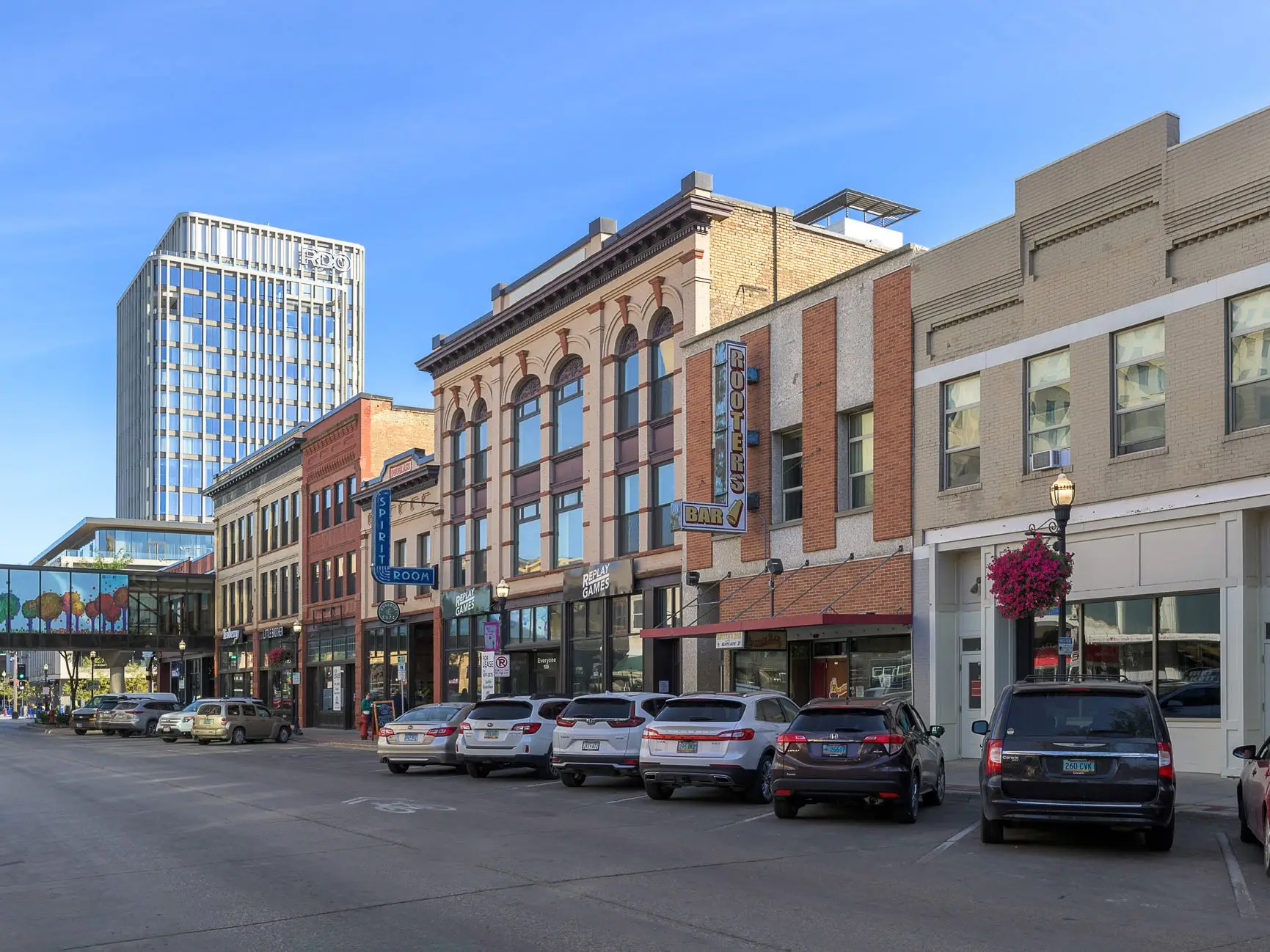 Parked cars line a downtown street with historic storefronts, a skyway, and a modern high-rise under blue sky.