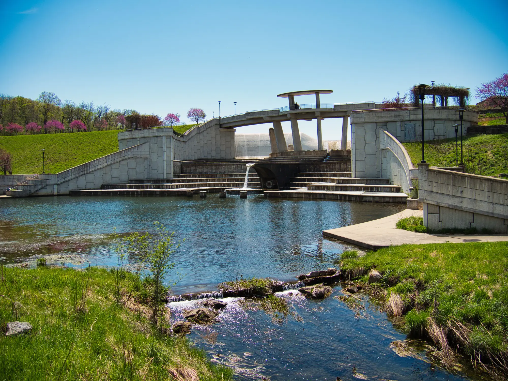 Terraced concrete spillway and pedestrian bridge overlook a pond in a landscaped park under a clear blue sky.
