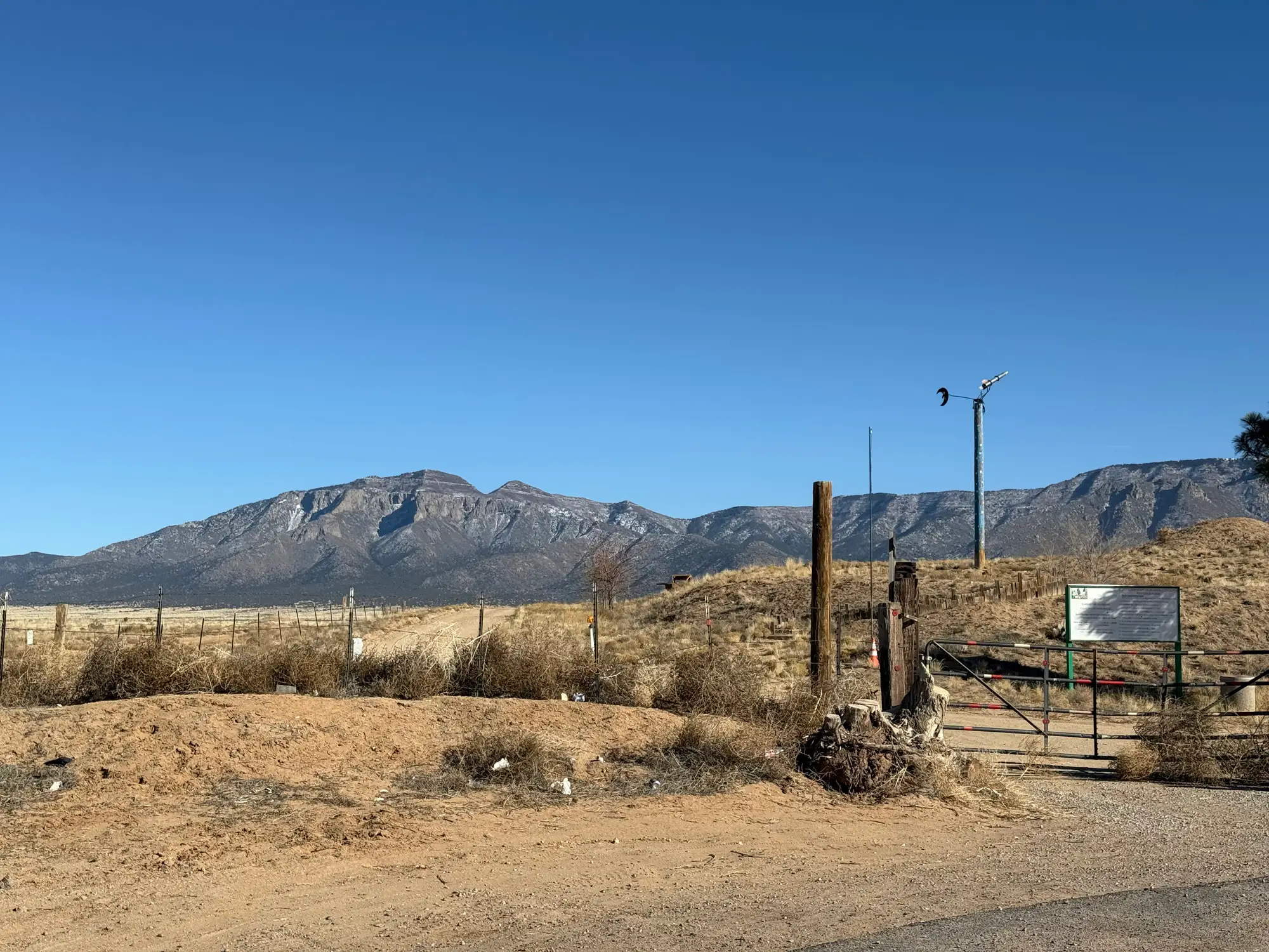 Desert roadside gate and fence stand before a dry hillside and rugged mountains under a clear blue sky.