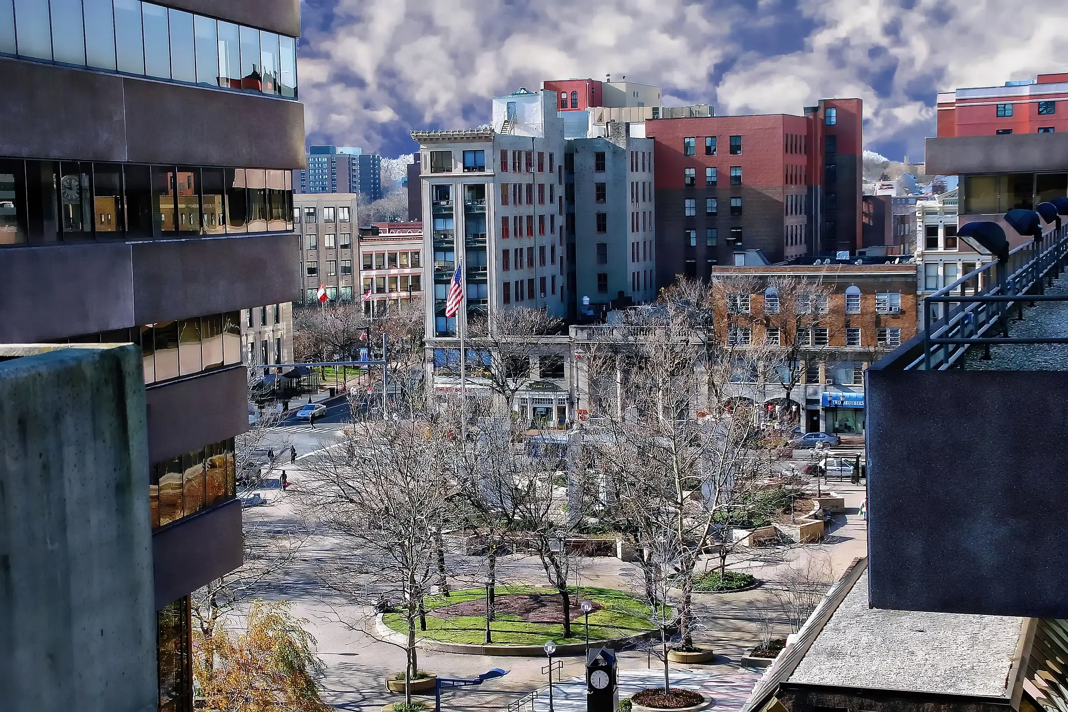 Elevated view of an urban plaza with leafless trees, surrounding buildings, and an American flag under a cloudy sky.