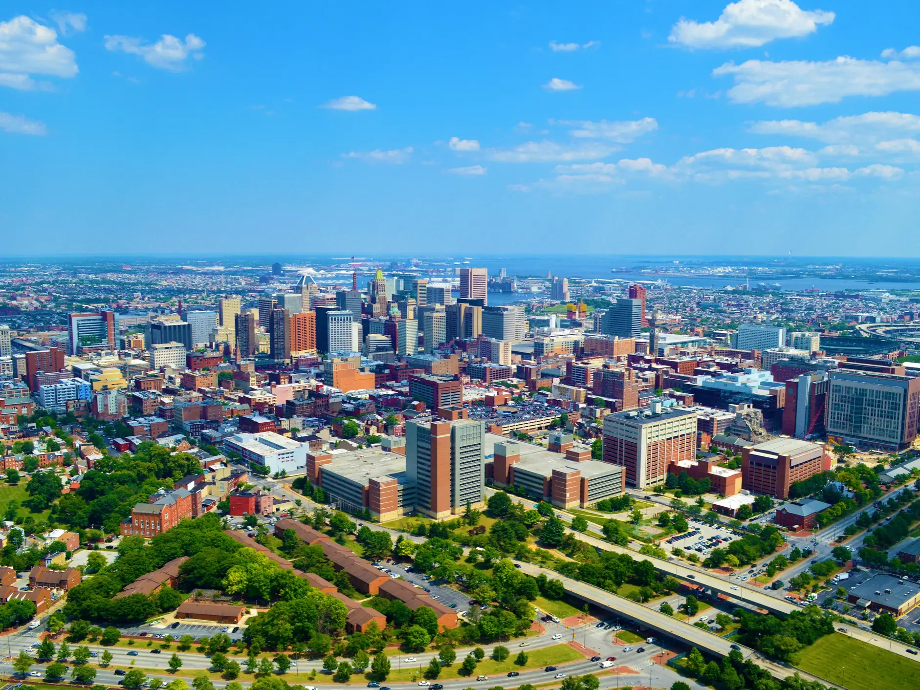 Aerial view of downtown Baltimore skyline with urban buildings, roads, greenery, and water under a partly cloudy sky.