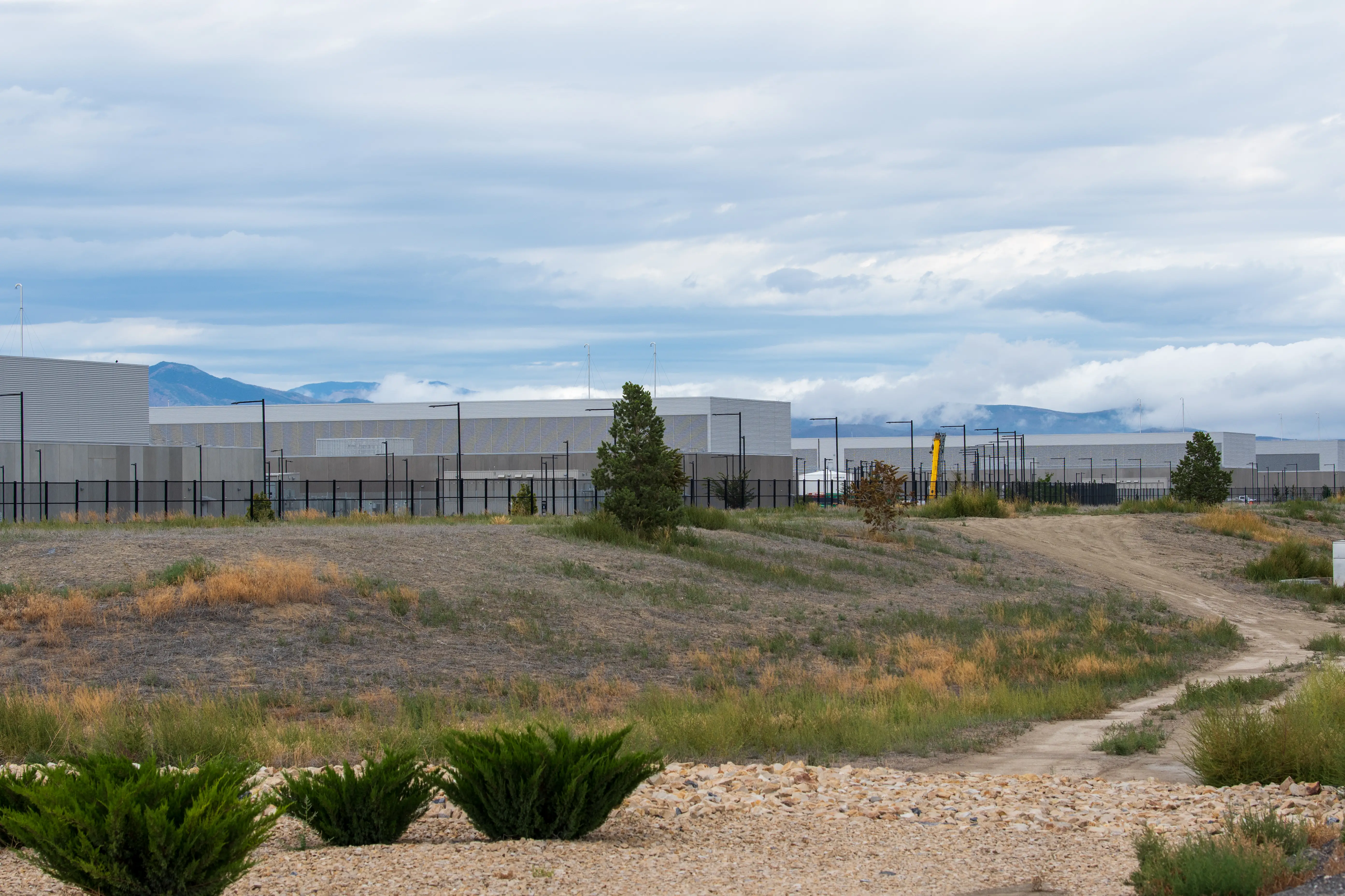 Fenced industrial buildings sit beyond a dirt road and scrubland under a cloudy mountain skyline.