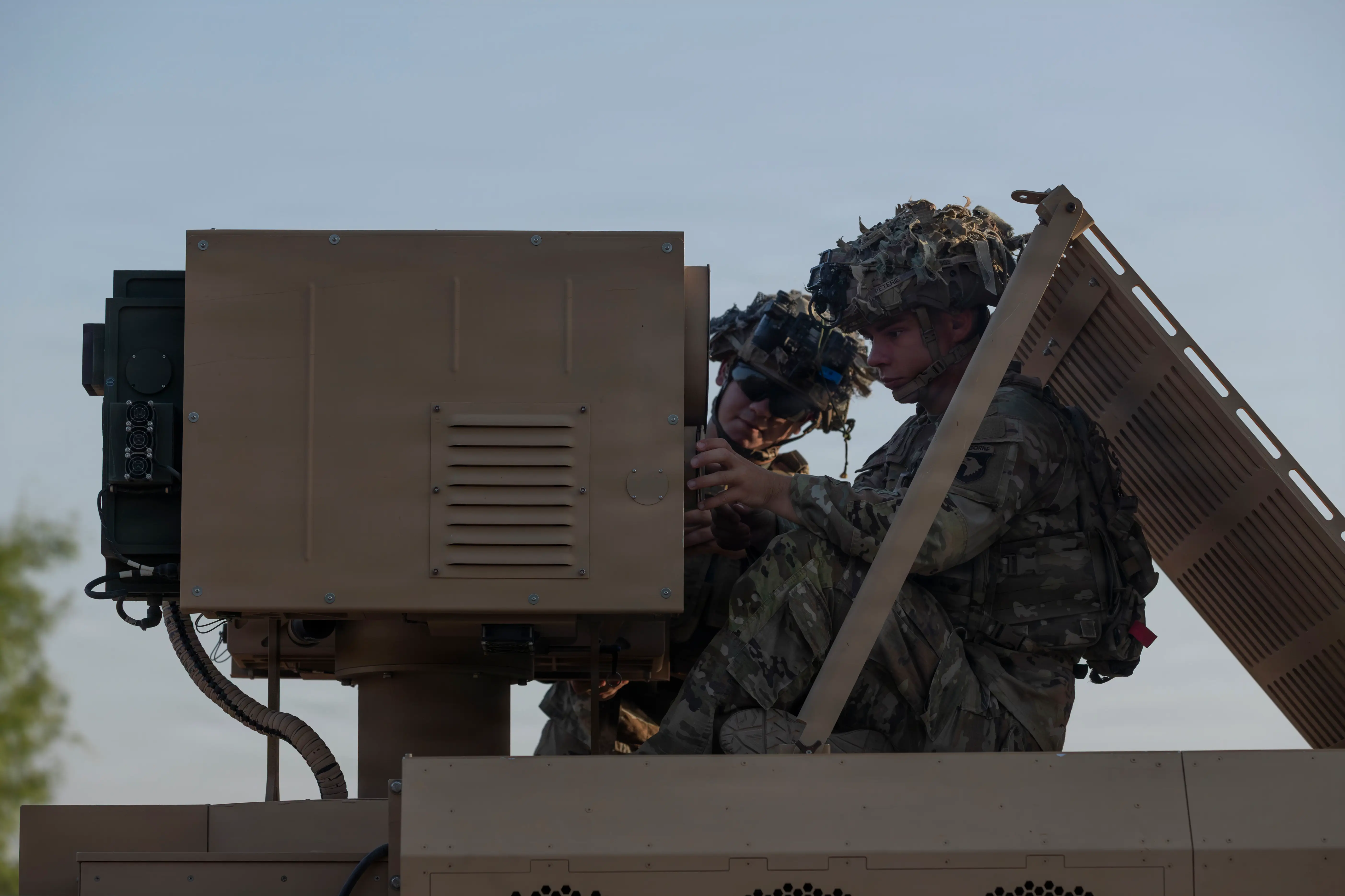 Two US Army soldiers sit next to an Army Multi-Purpose High Energy Laser weapon mounted on a vehicle.