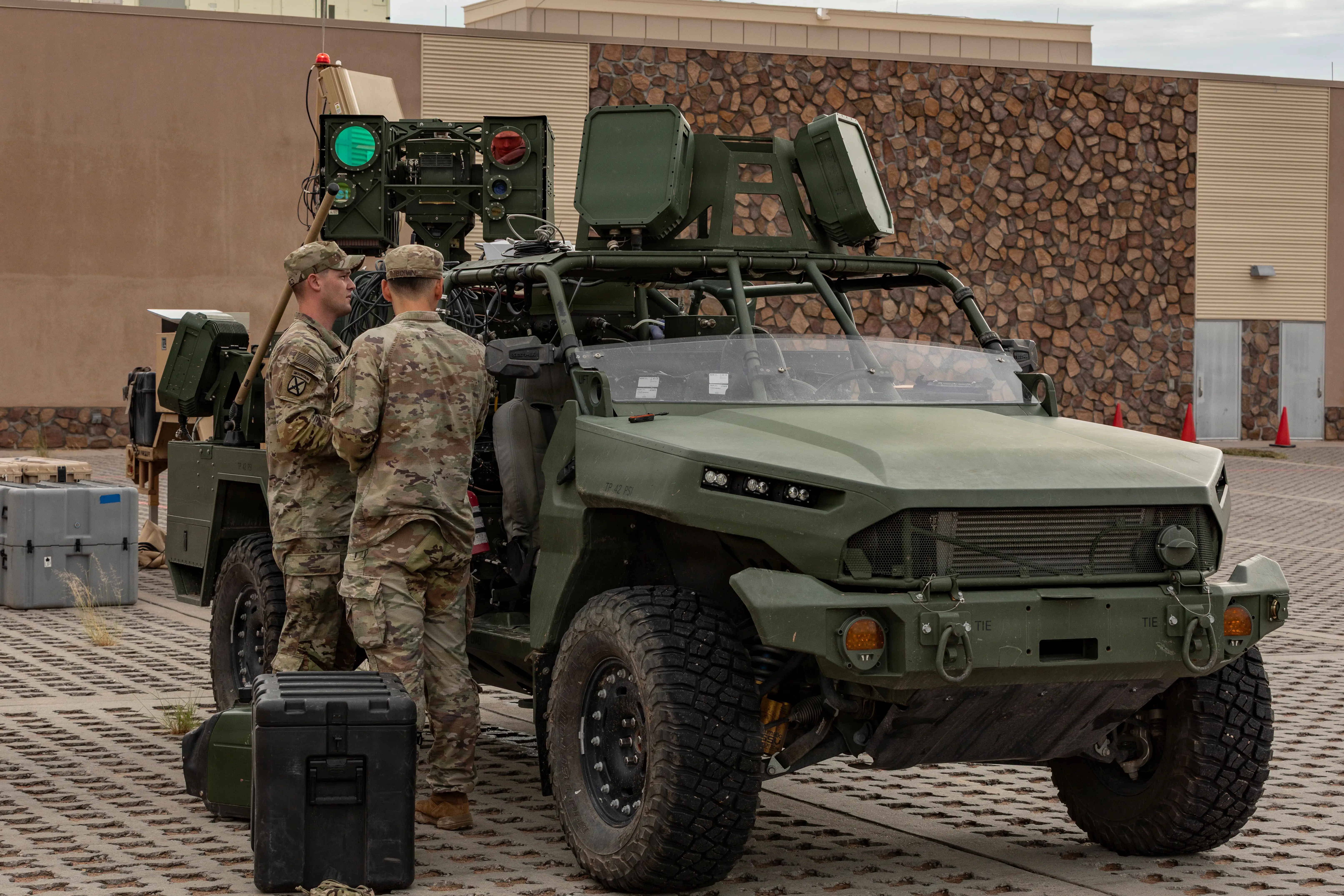 US Army soldiers stand near a vehicle that has laser weapons mounted on top of it.