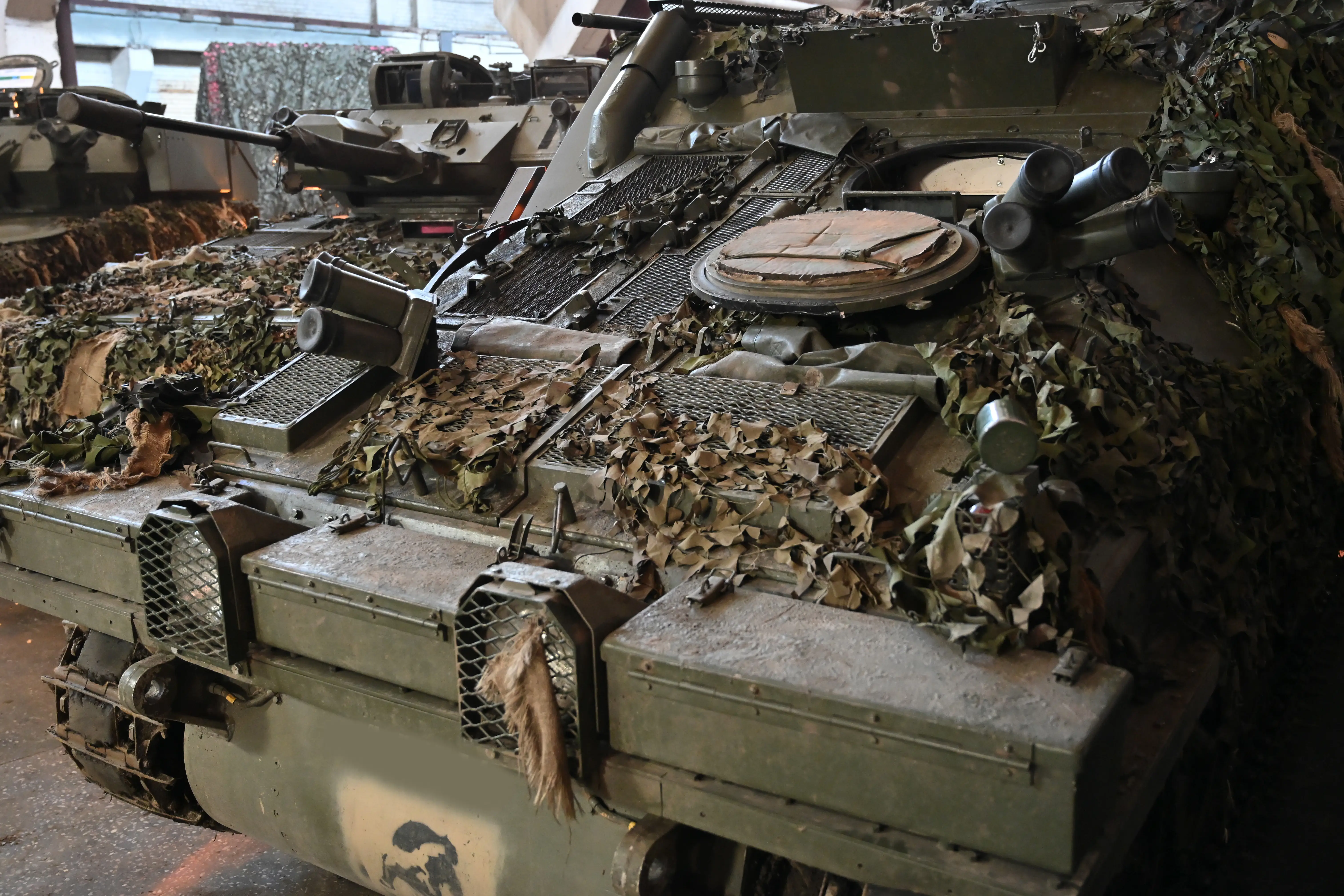 Armored military vehicles with camouflage netting are parked closely together in an indoor display area.
