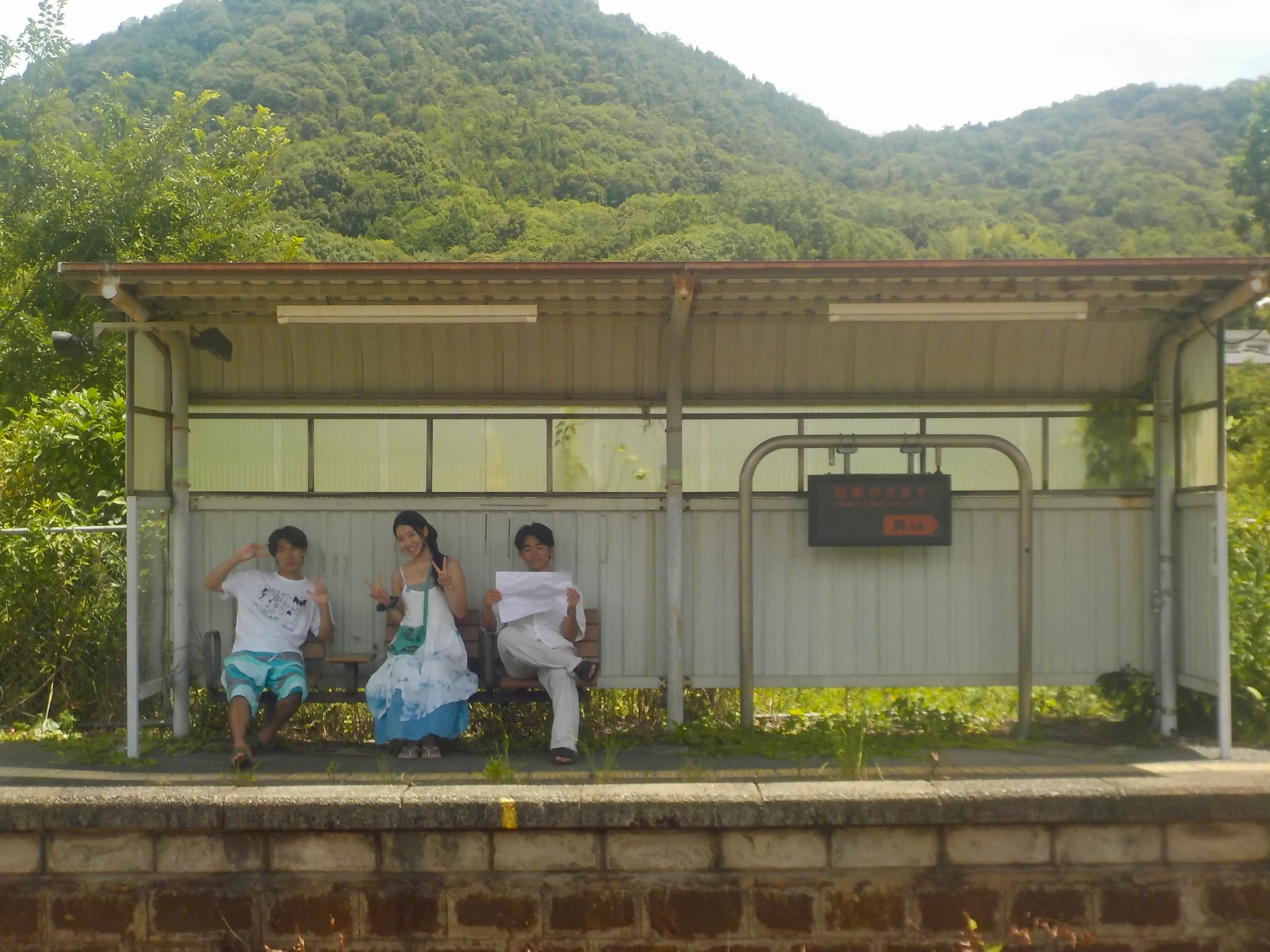 Three people sit on a rural train platform bench beneath a shelter with forested hills behind.