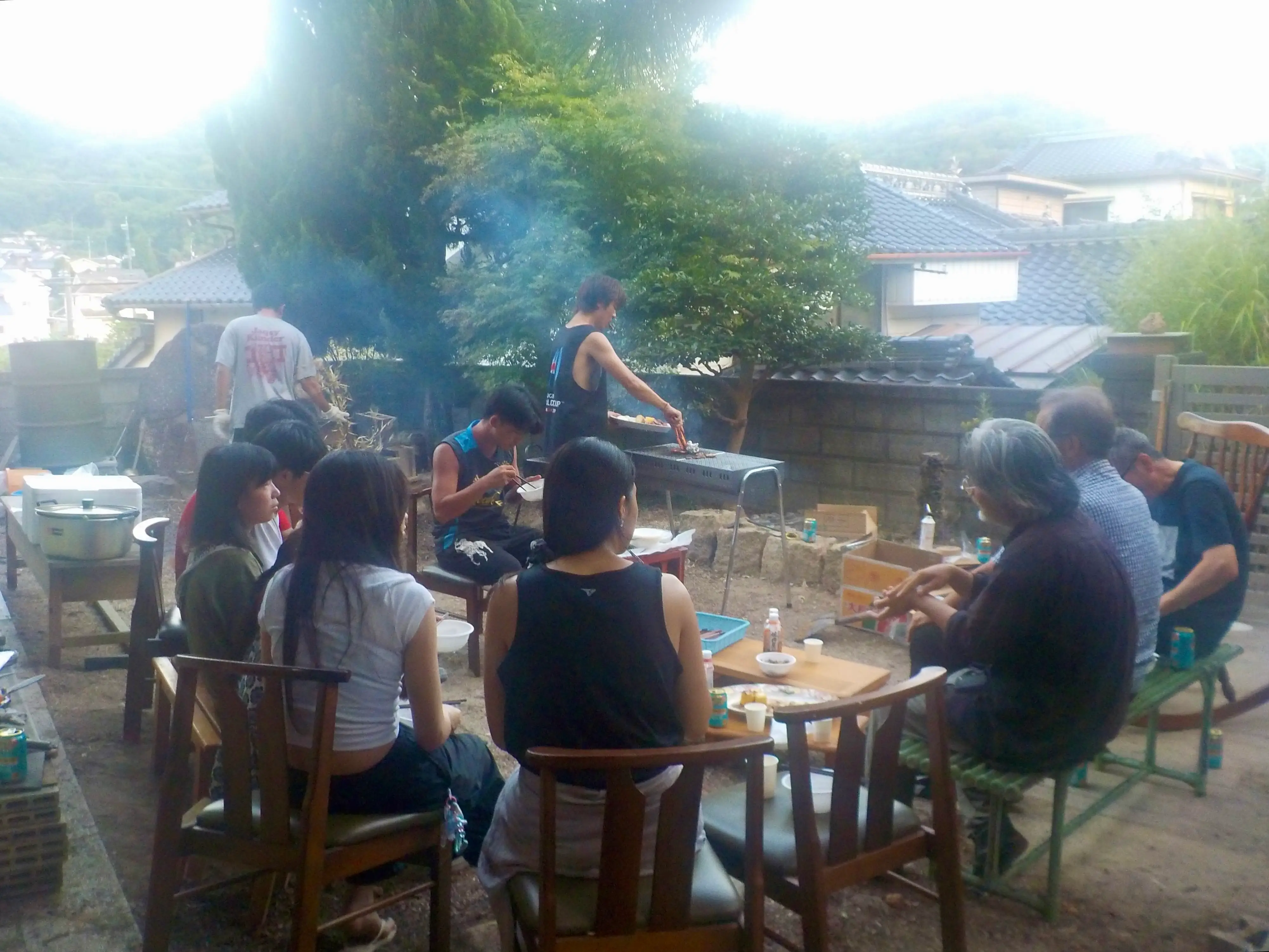 Group gathers around outdoor tables while food cooks on a grill in a residential garden setting.