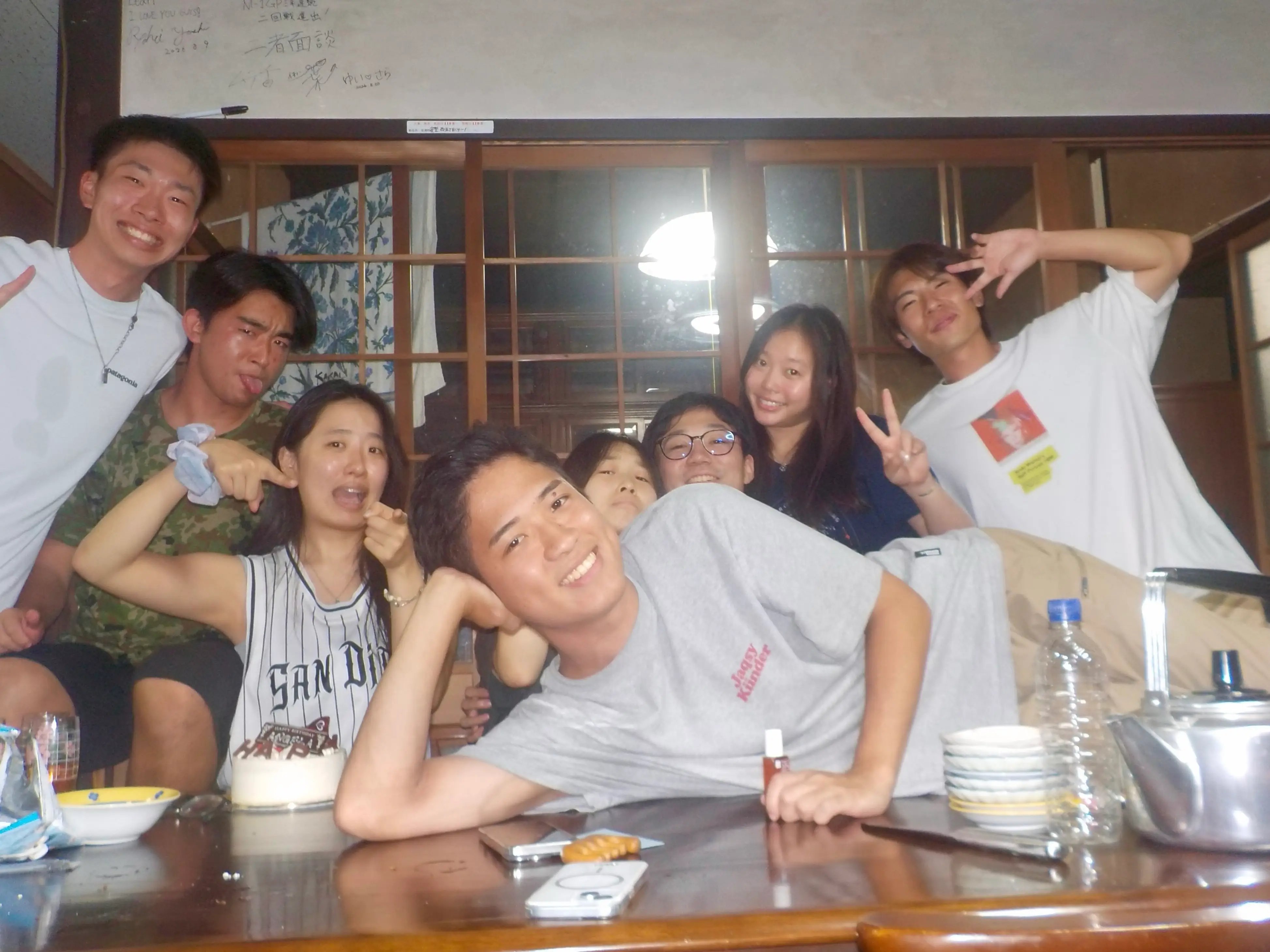 Eight people pose playfully around a dining table in a wood-paneled room with sliding glass doors.