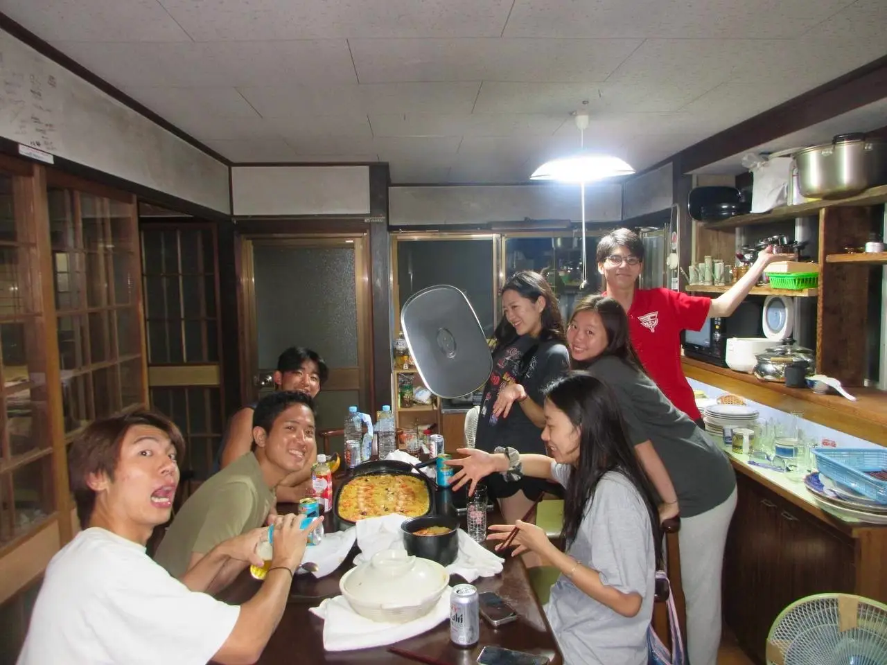 Seven people gather around a dining table with cookware and drinks in a home kitchen.