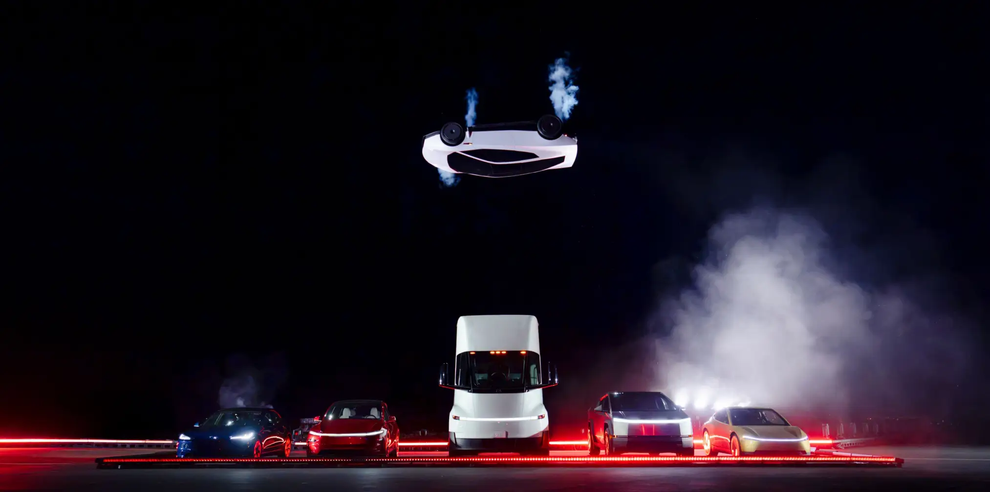 A Tesla Roadster prototype flips in the air over a line of other Tesla vehicles.