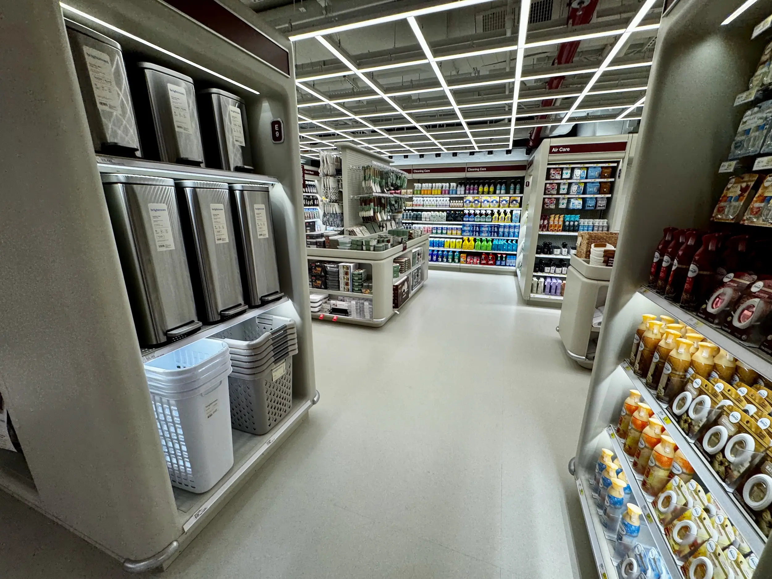 Bright retail aisle displays trash bins, laundry baskets, cleaning supplies, car care products, and bottled household goods.