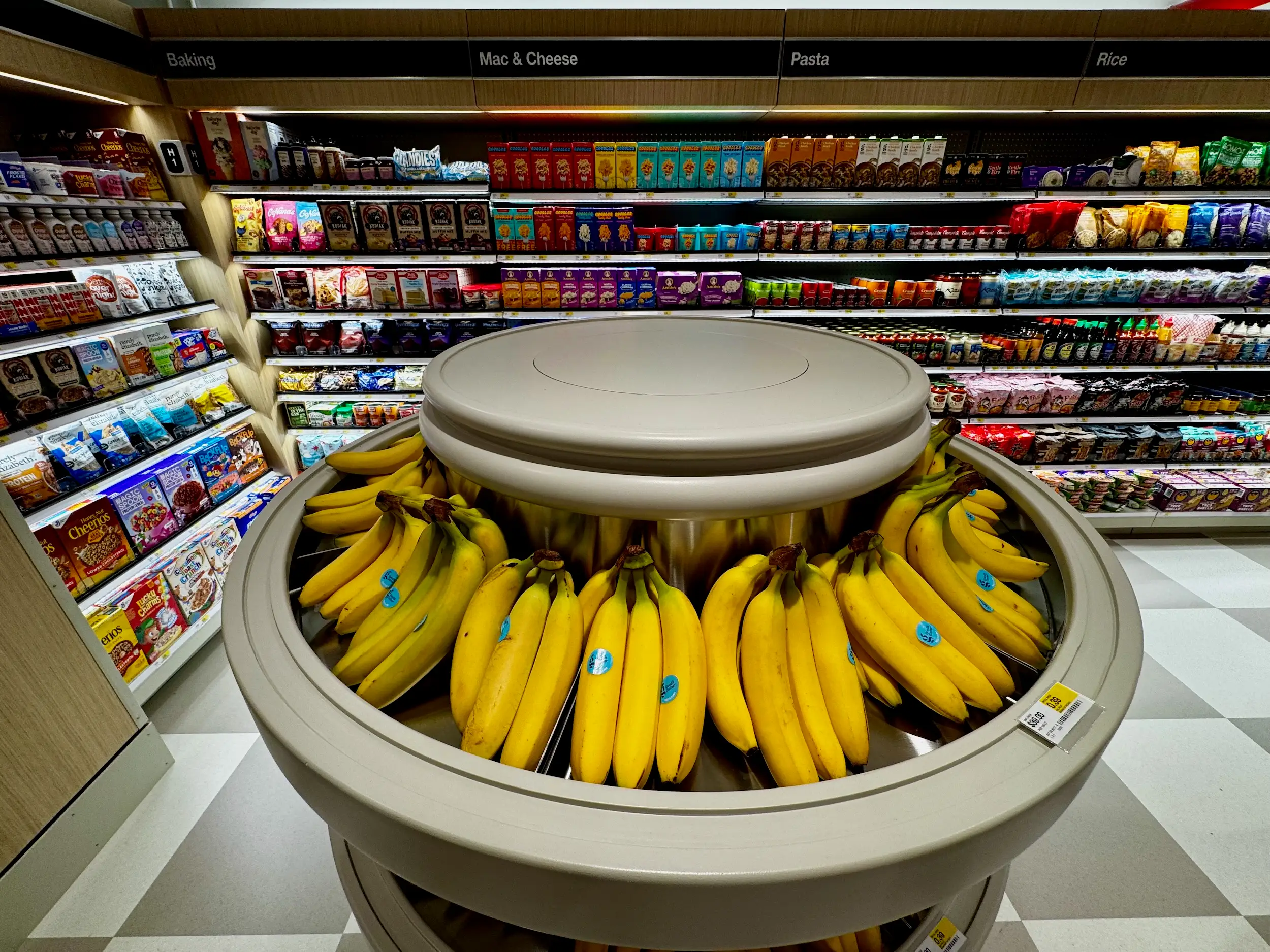 Bananas displayed on a circular produce stand in a grocery aisle with packaged foods and overhead category signs.