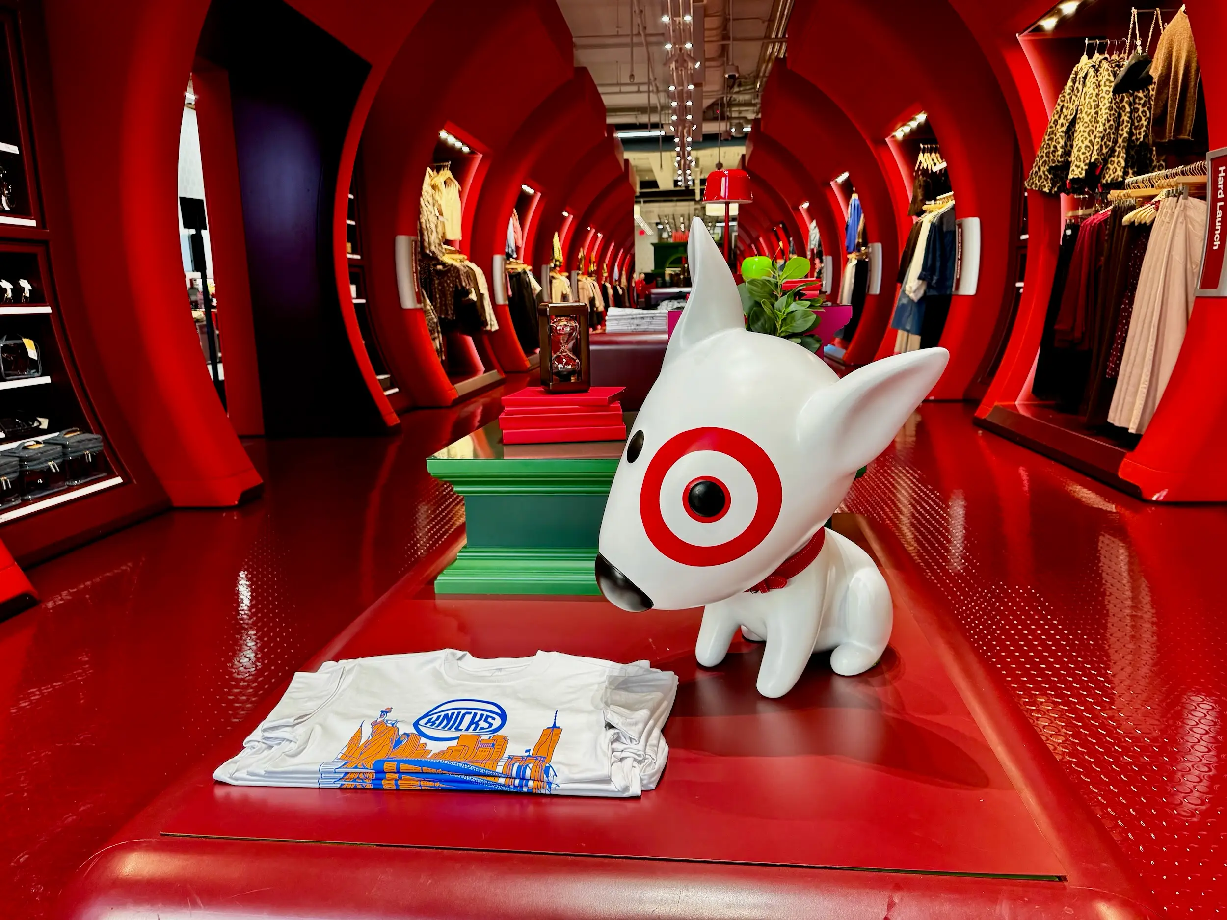 Target retail aisle with red arch displays, Bullseye dog statue, clothing racks, and folded graphic T-shirt.