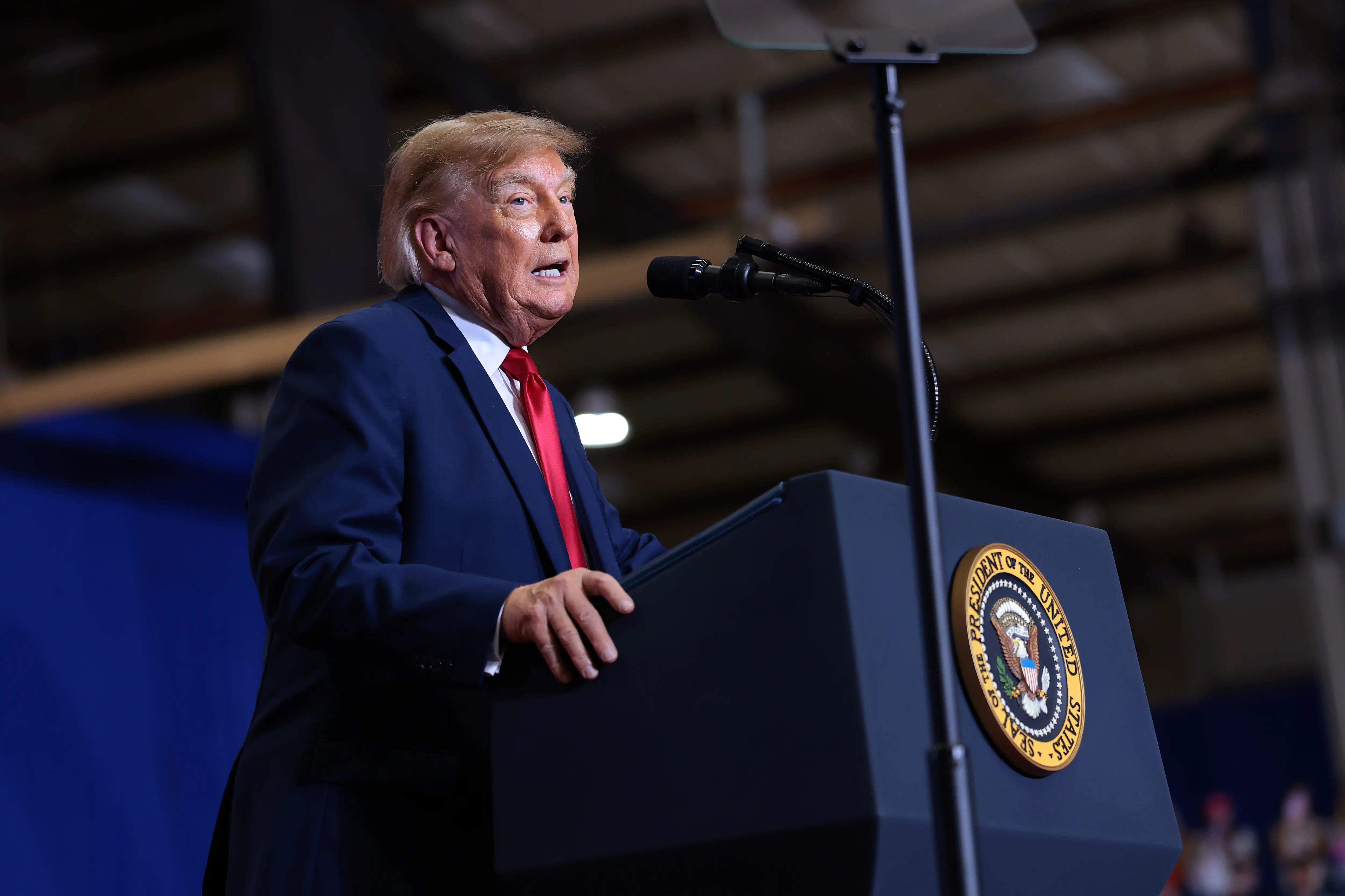 Donald Trump speaks at a podium with the presidential seal in an indoor venue.