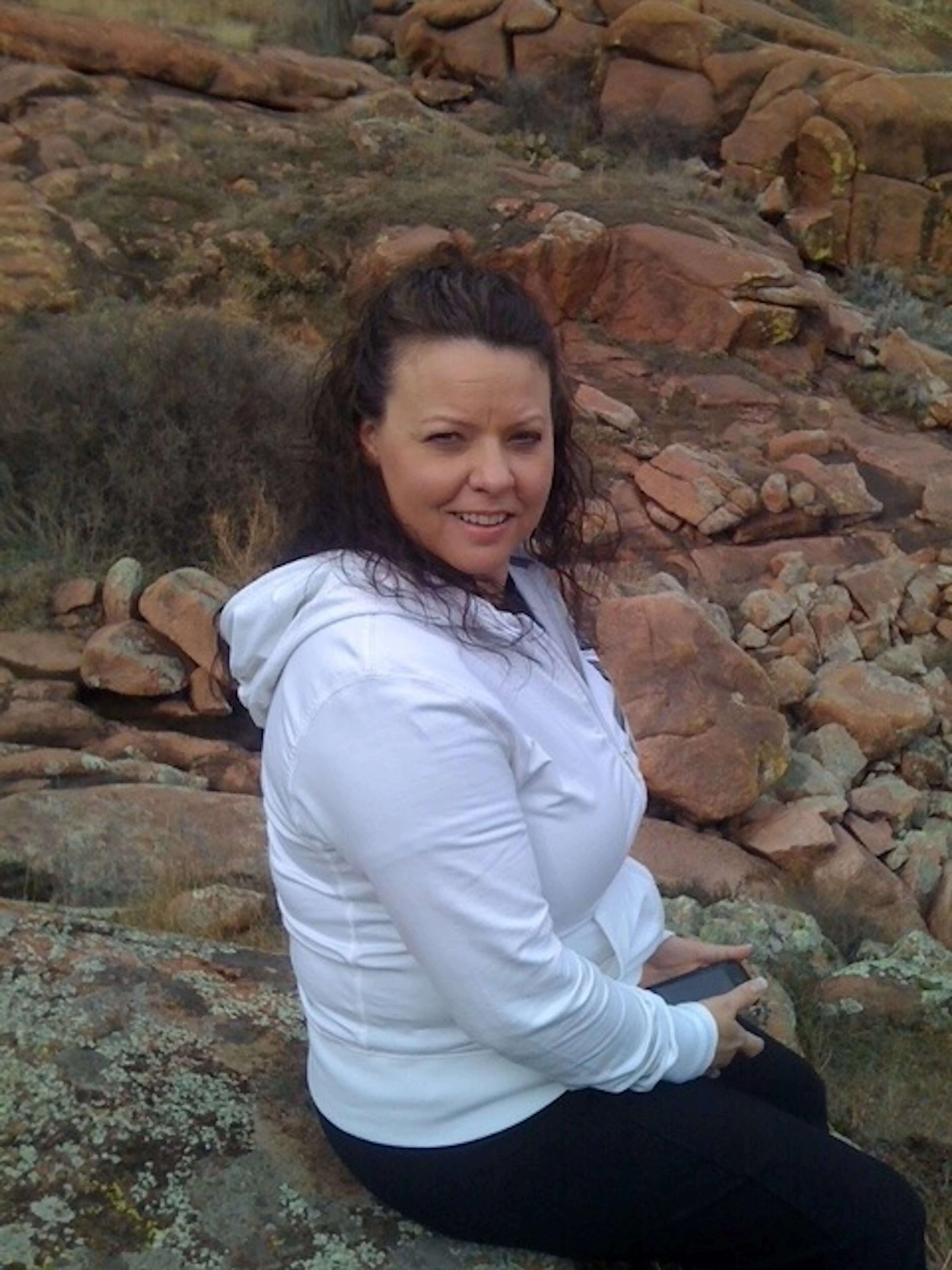Person in a white hoodie sits on rocky terrain holding a phone in an outdoor landscape.