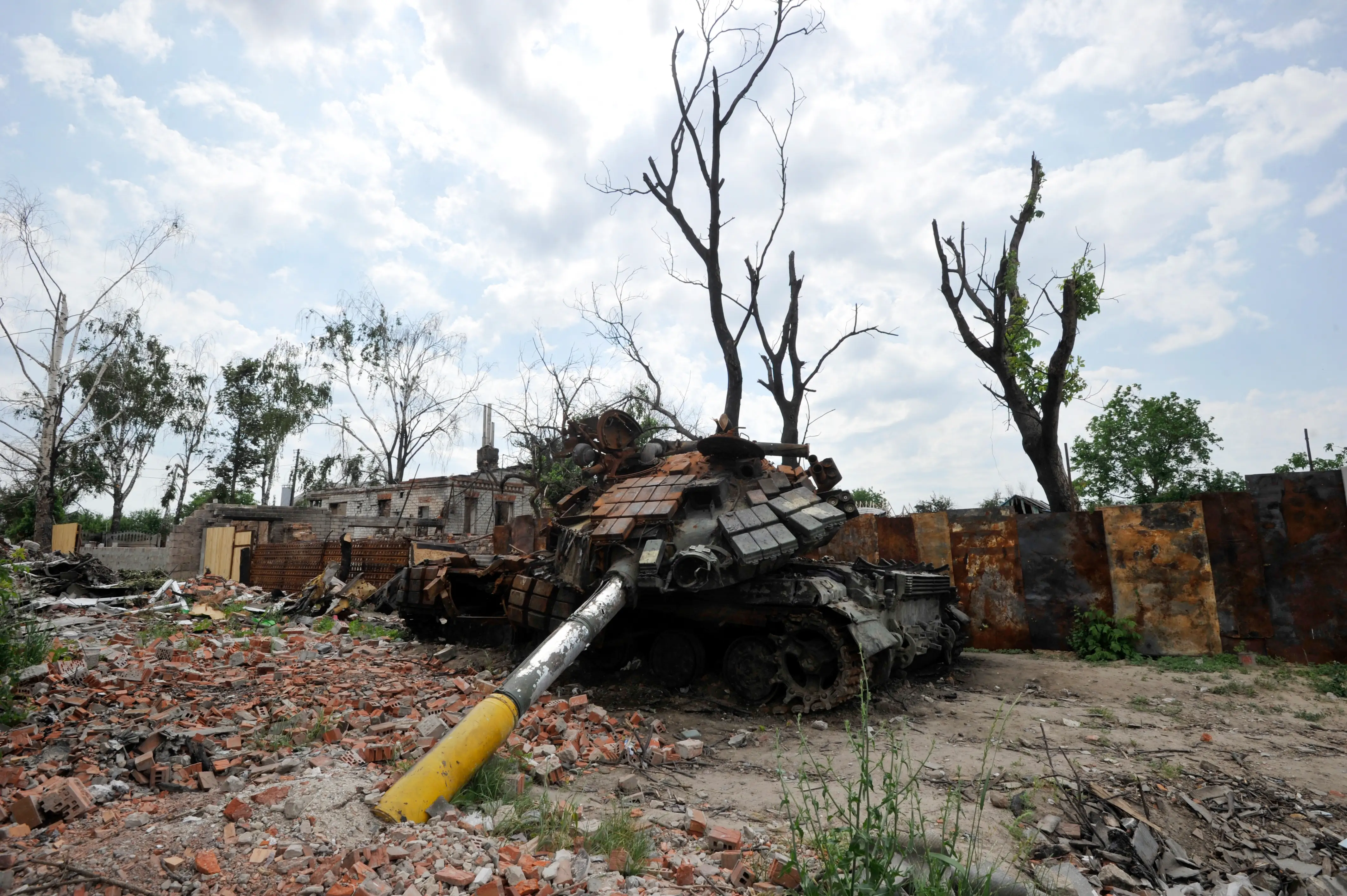 Destroyed armored tank lies amid rubble and damaged trees beside ruined walls under a cloudy sky.