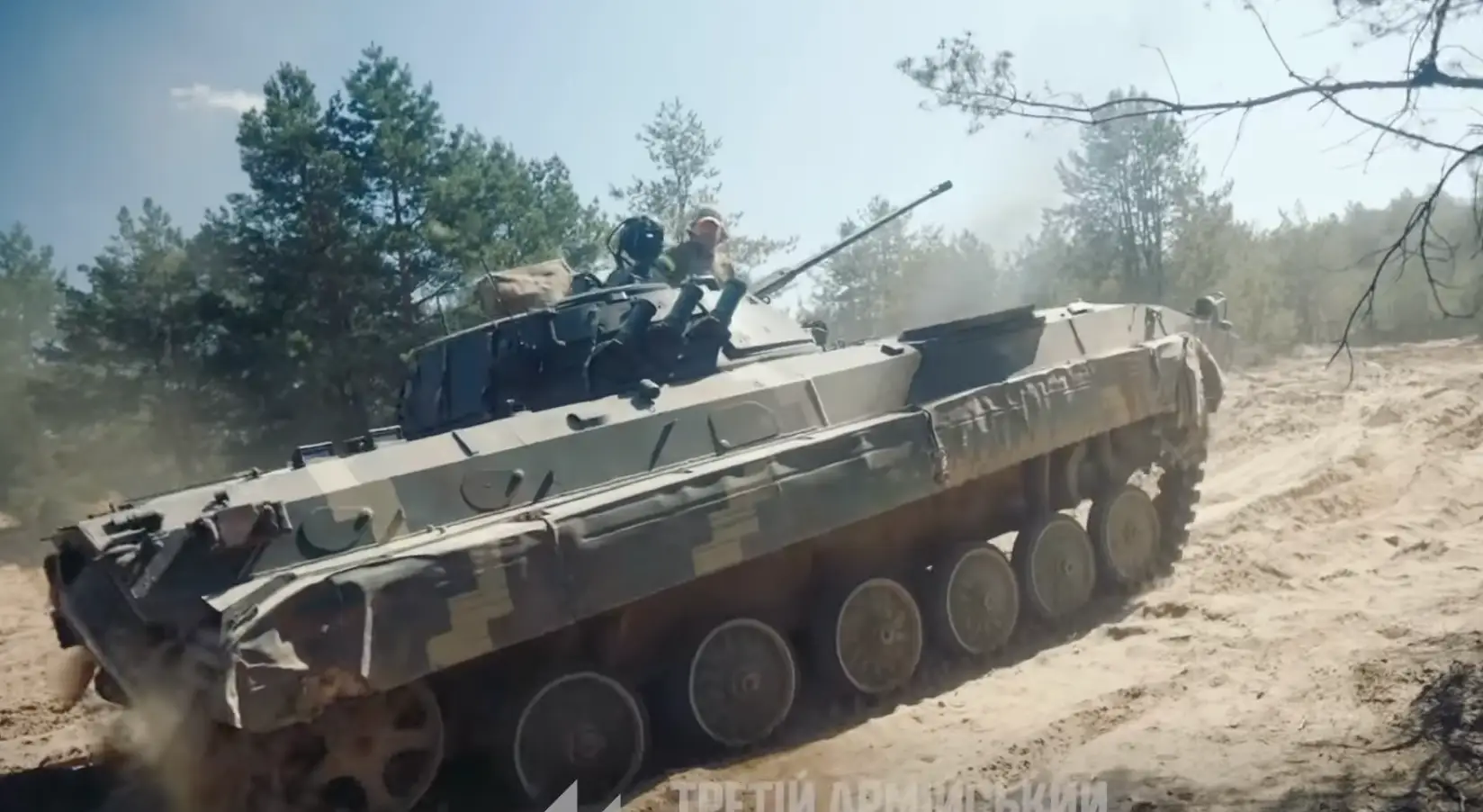 Tracked armored vehicle drives along a sandy forest road with two people riding on top.