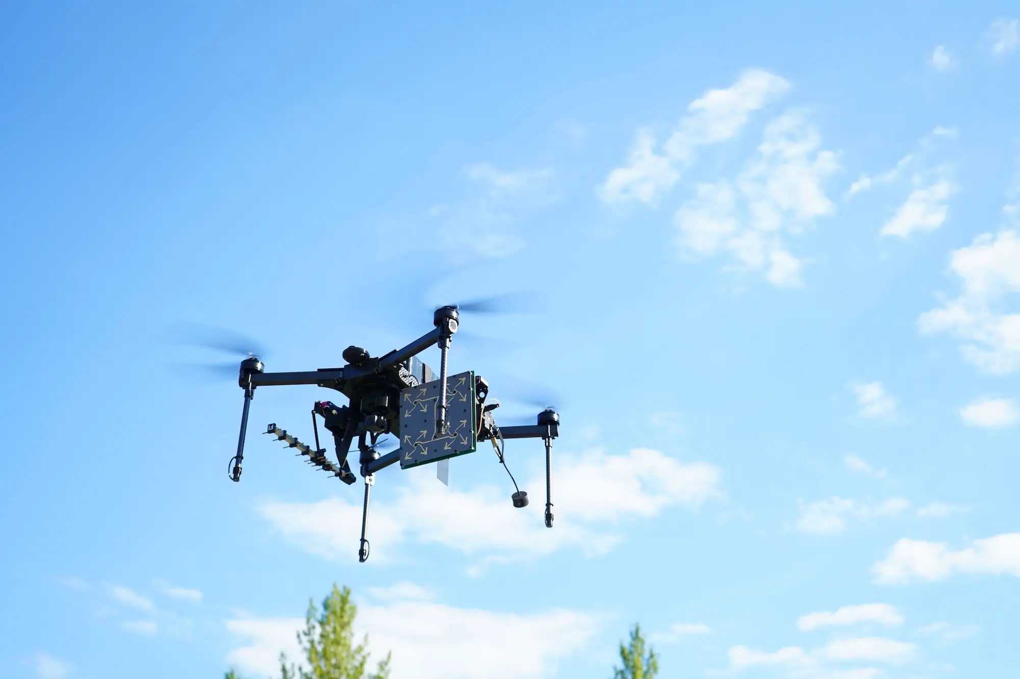 Multirotor drone flies against a blue sky carrying sensors and a patterned calibration panel.